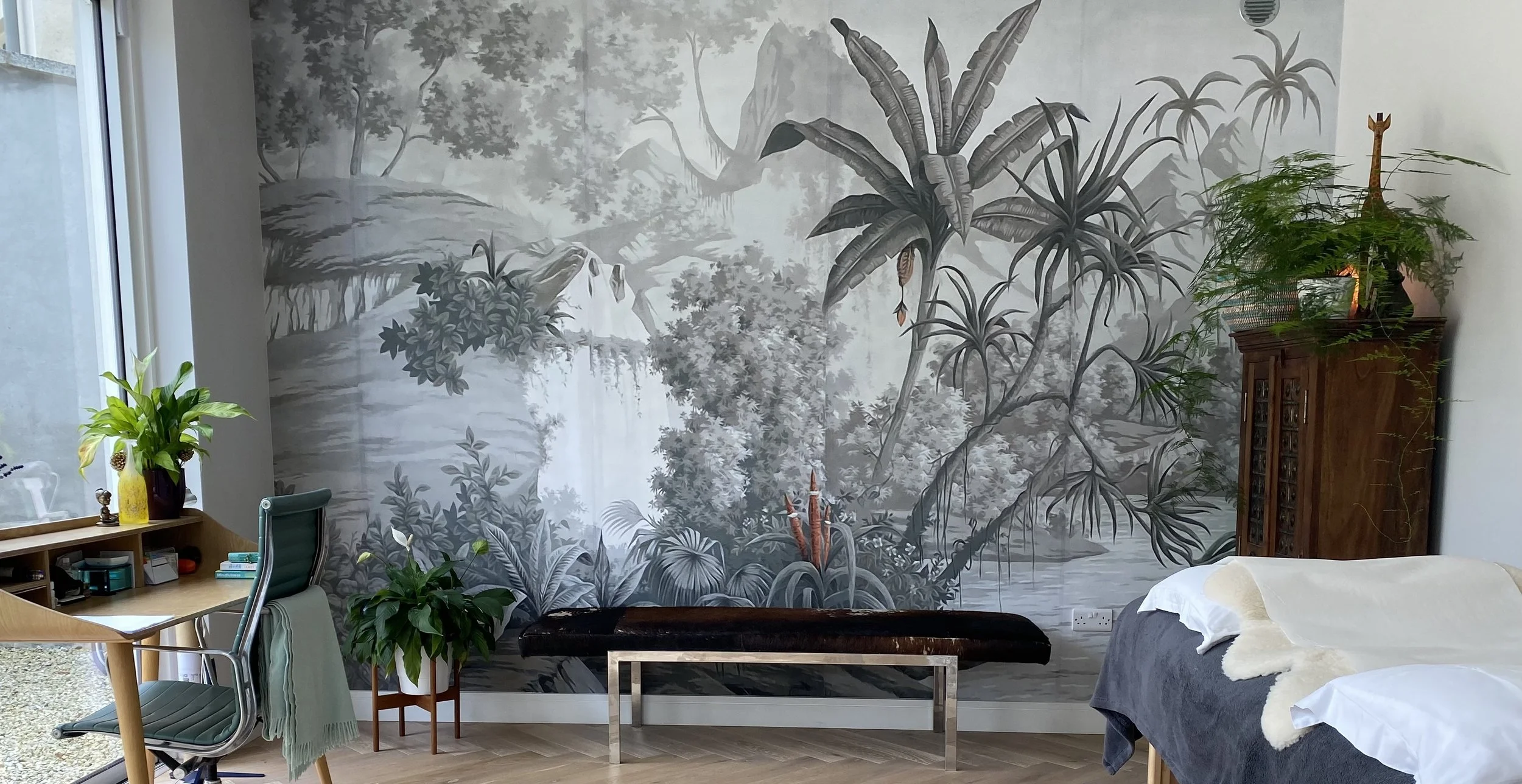 Living room with a black-and-white jungle-themed mural on the wall, featuring large tropical leaves and trees. There are various plants, a wooden cabinet with a plant on top, a bed with a white blanket, a desk with a yellow vase with green leaves, and a window to the left letting in natural light.