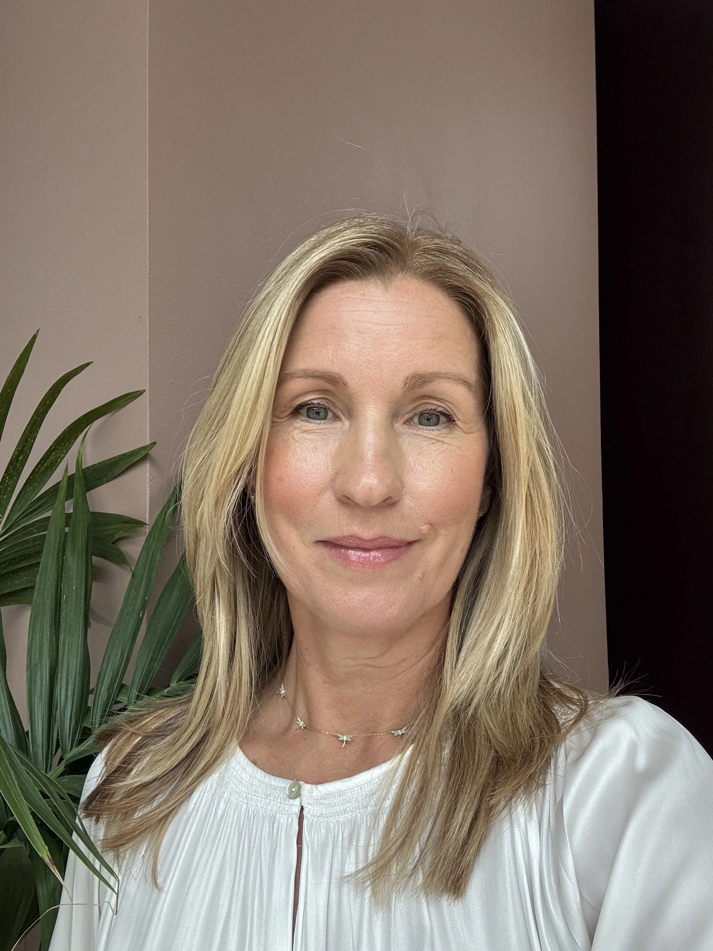 A smiling woman with blonde hair wearing a white blouse, standing next to a green plant indoors.