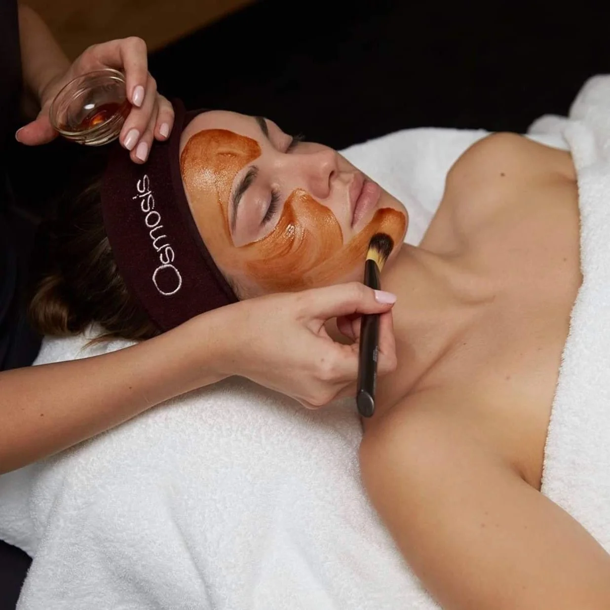 A woman lying on her back with a white towel around her chest, receiving a facial treatment with a facial mask applied on her face. An esthetician is using a brush to apply a product near her collarbone, and holding a small glass bowl with facial products. The woman is wearing a headband that says 'Ossuos'.