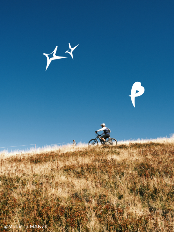Une personne fait du vélo en montée dans un champ de herbes sèches sous un ciel bleu clair, avec quelques formes graphiques blanches dans le ciel.