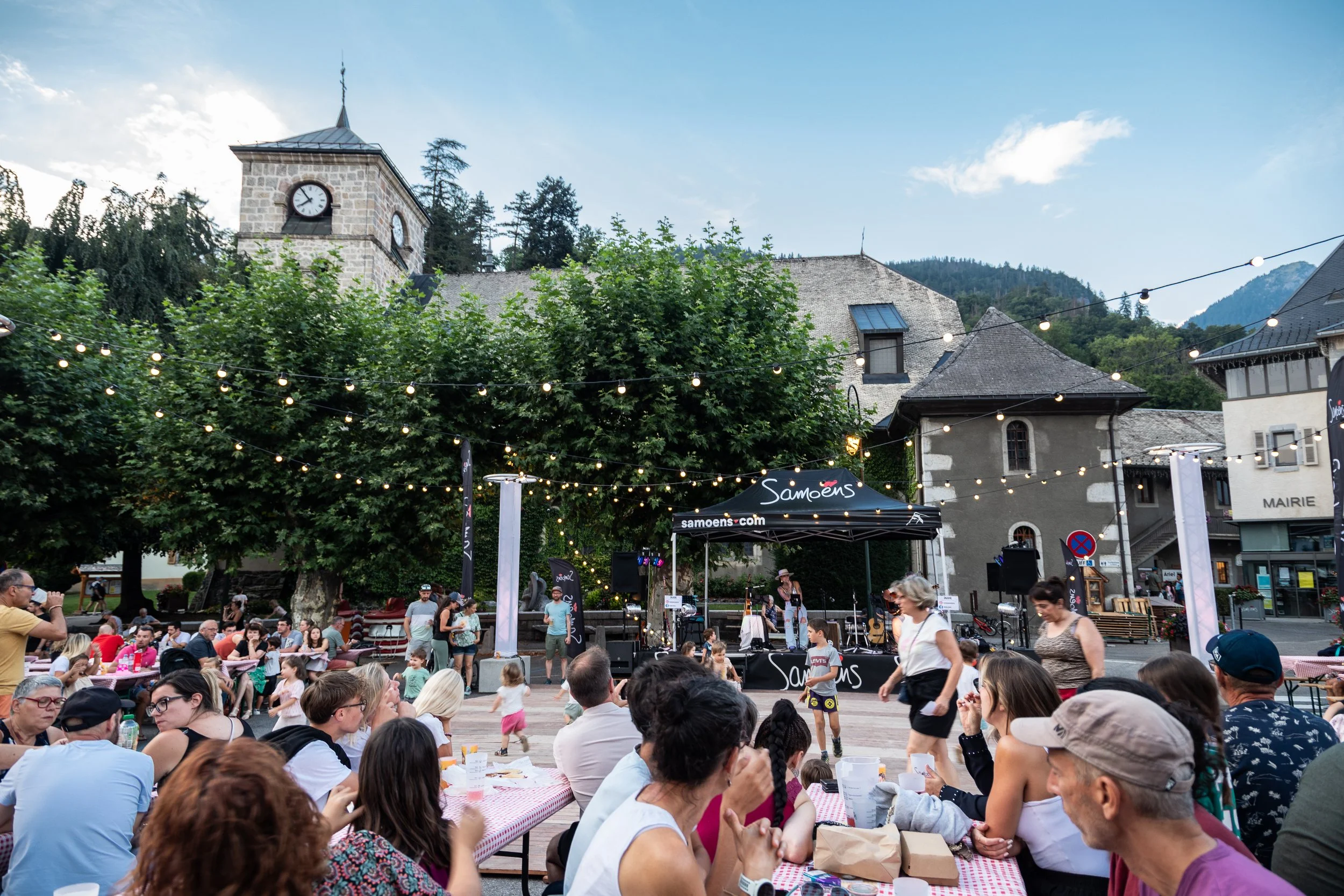 Une scène de fête en plein air avec des gens assis autour de tables, un groupe de musiciens sur une scène avec un parasol noir marqué « Samoëns », des arbres et des bâtiments anciens en arrière-plan, des guirlandes lumineuses suspendues dans l'air.