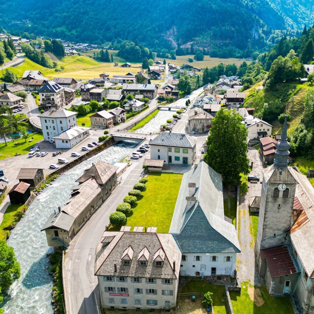 Vue aérienne d'un village alpin avec une rivière traversant la centre, entouré de maisons, d'arbres, et de montagnes dans le fond.