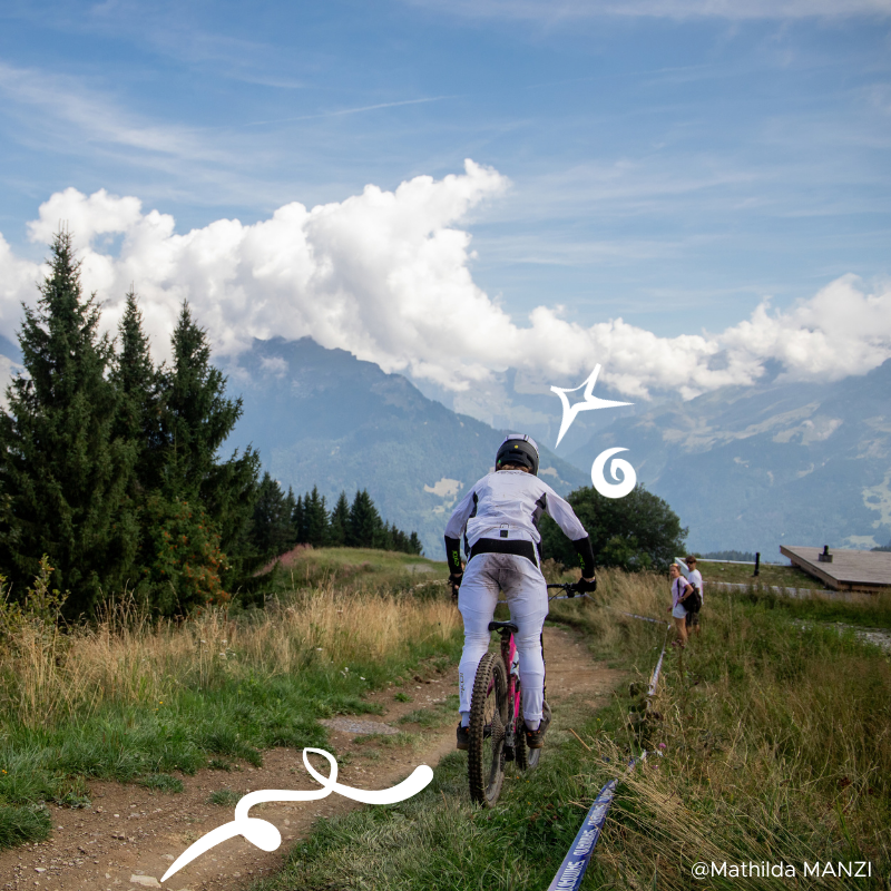 Une personne en tenue de cyclisme descend une piste de montagne à VTT, avec un paysage de montagnes, de forêt et un ciel en arrière-plan, quelques spectateurs observant la scène.