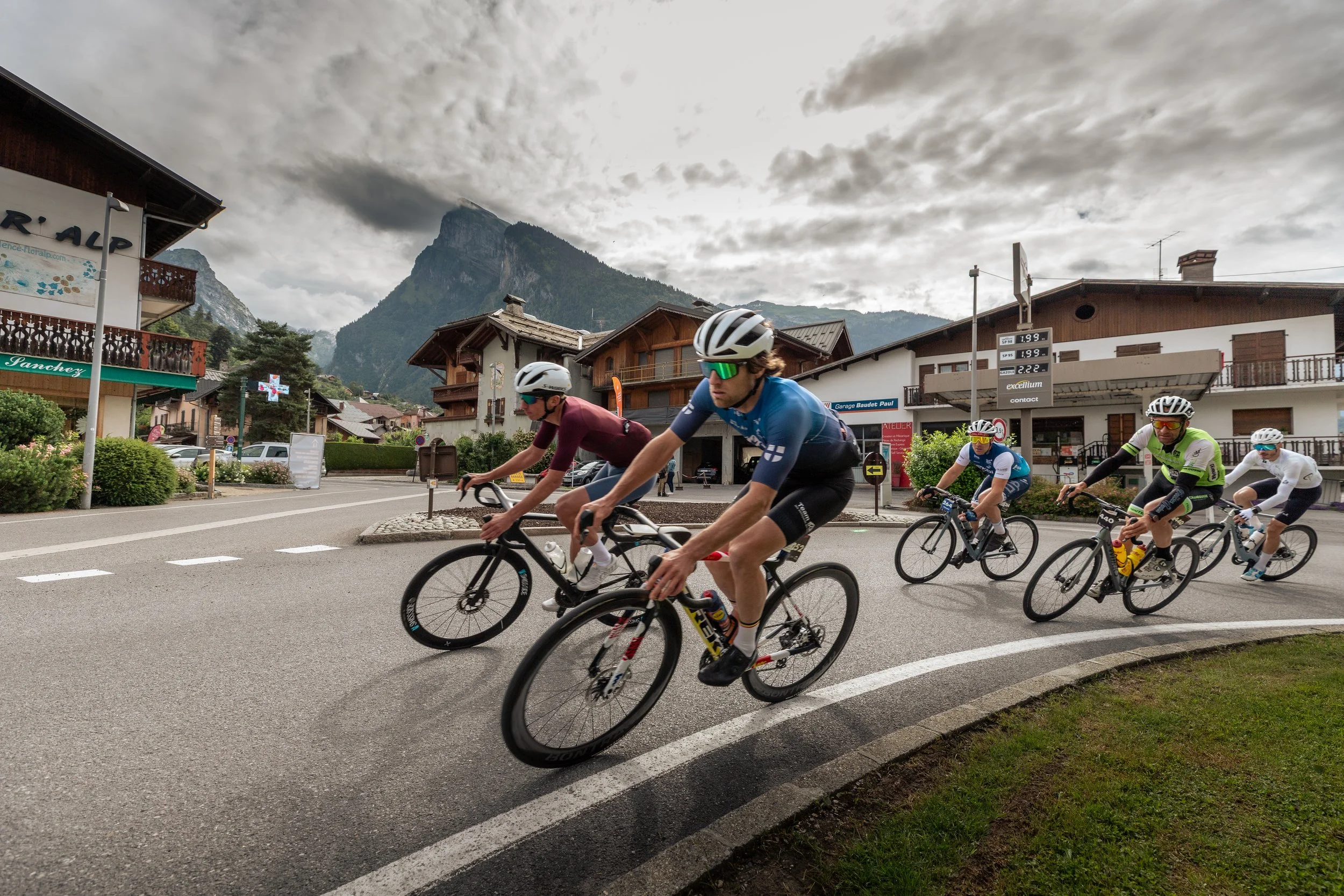 Groupe de cyclistes participant à une course dans un village alpin avec des montagnes en arrière-plan et une météo nuageuse.