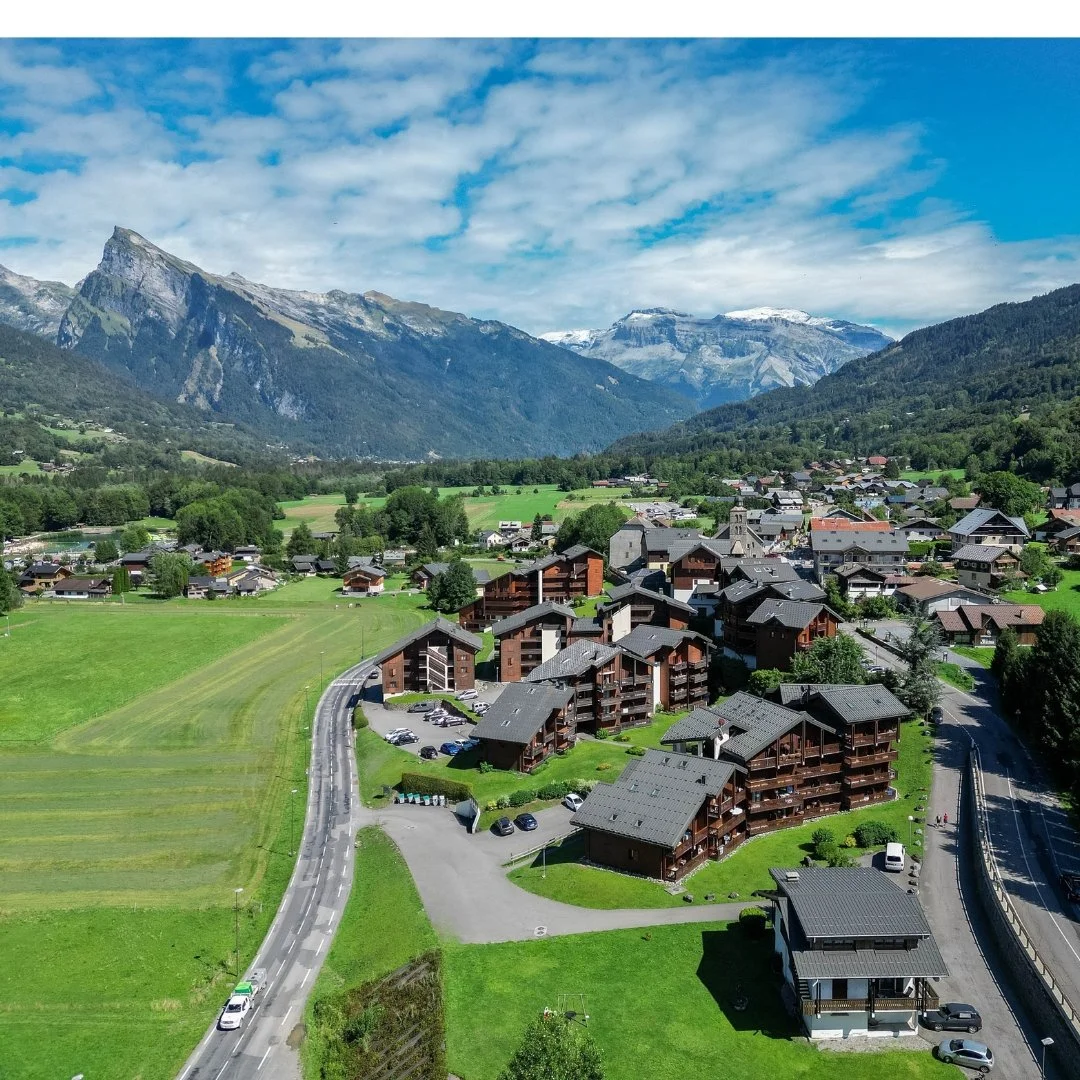 Vallée alpine avec des maisons en bois, montagnes en arrière-plan, et un ciel nuageux.