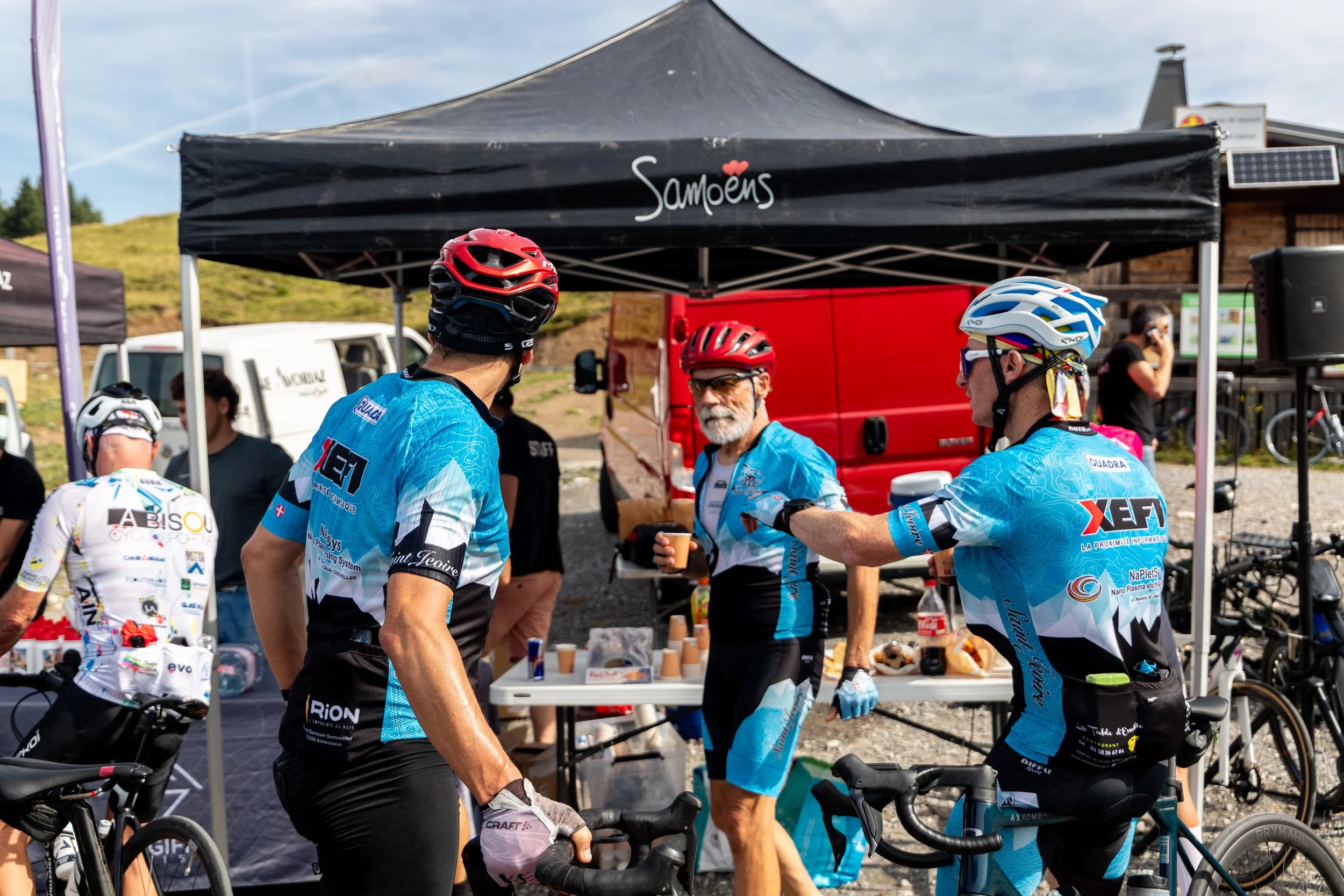 Groupe de cyclistes en tenue de cyclisme buvant et discutant sous une tente lors d'un événement en plein air.