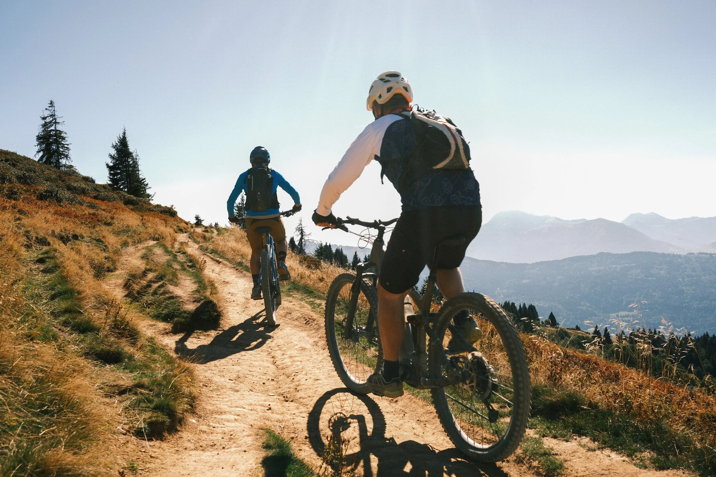 Deux personnes font du vélo sur un sentier de montagne, portant des casques et des sacs à dos, avec des montagnes en arrière-plan.