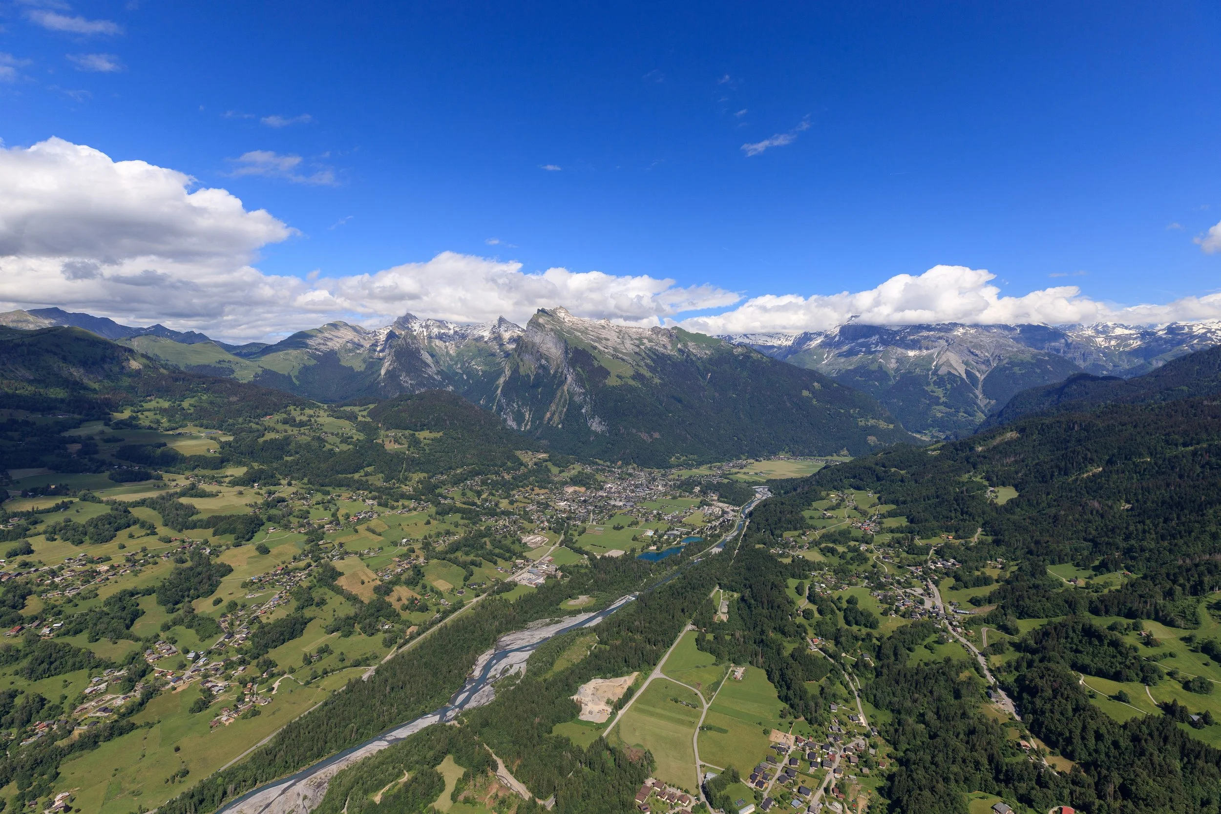 Paysage de montagne avec des sommets enneigés, des vallées verdoyantes, un fleuve serpentant à travers la vallée et un village rural au premier plan, sous un ciel bleu partiellement nuageux.