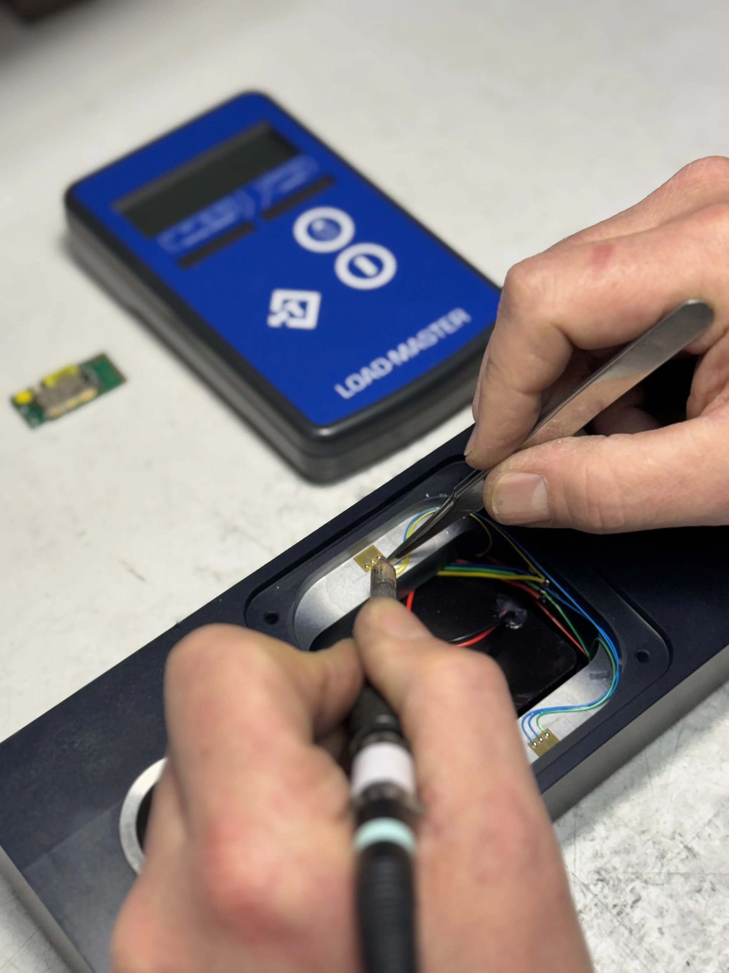 Close-up of hands working inside an electronic device with colorful wires, a small circuit board nearby, and a blue digital load tester in the background.