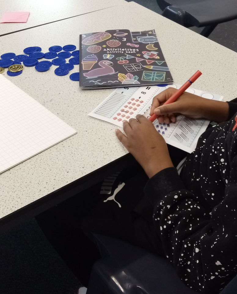 A child's hand writing with an orange marker on a worksheet with colorful patterns. On the desk, there is a black activity book with colorful illustrations, a set of blue tokens, and a notebook.