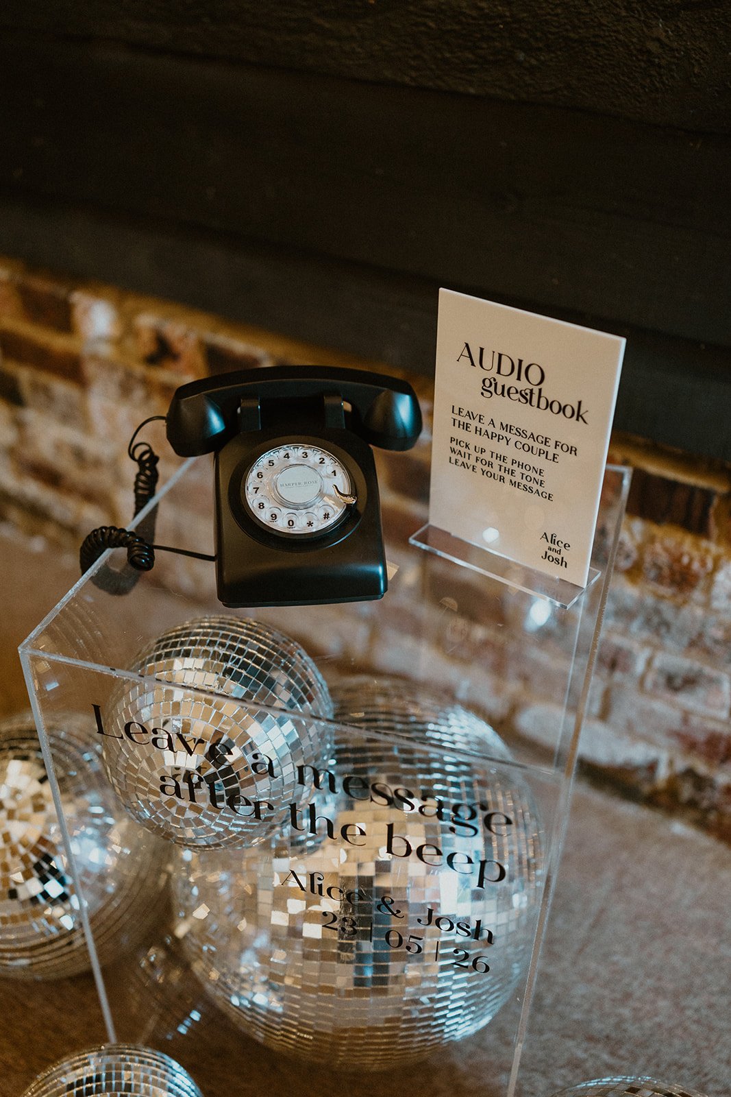 A vintage black rotary phone on a clear acrylic stand with a sign that reads 'Audio Guestbook' and instructions to leave a message for the couple, Alice and Josh, with a date of 23-05-26. In front, there are disco balls with a message that says, 'Lea