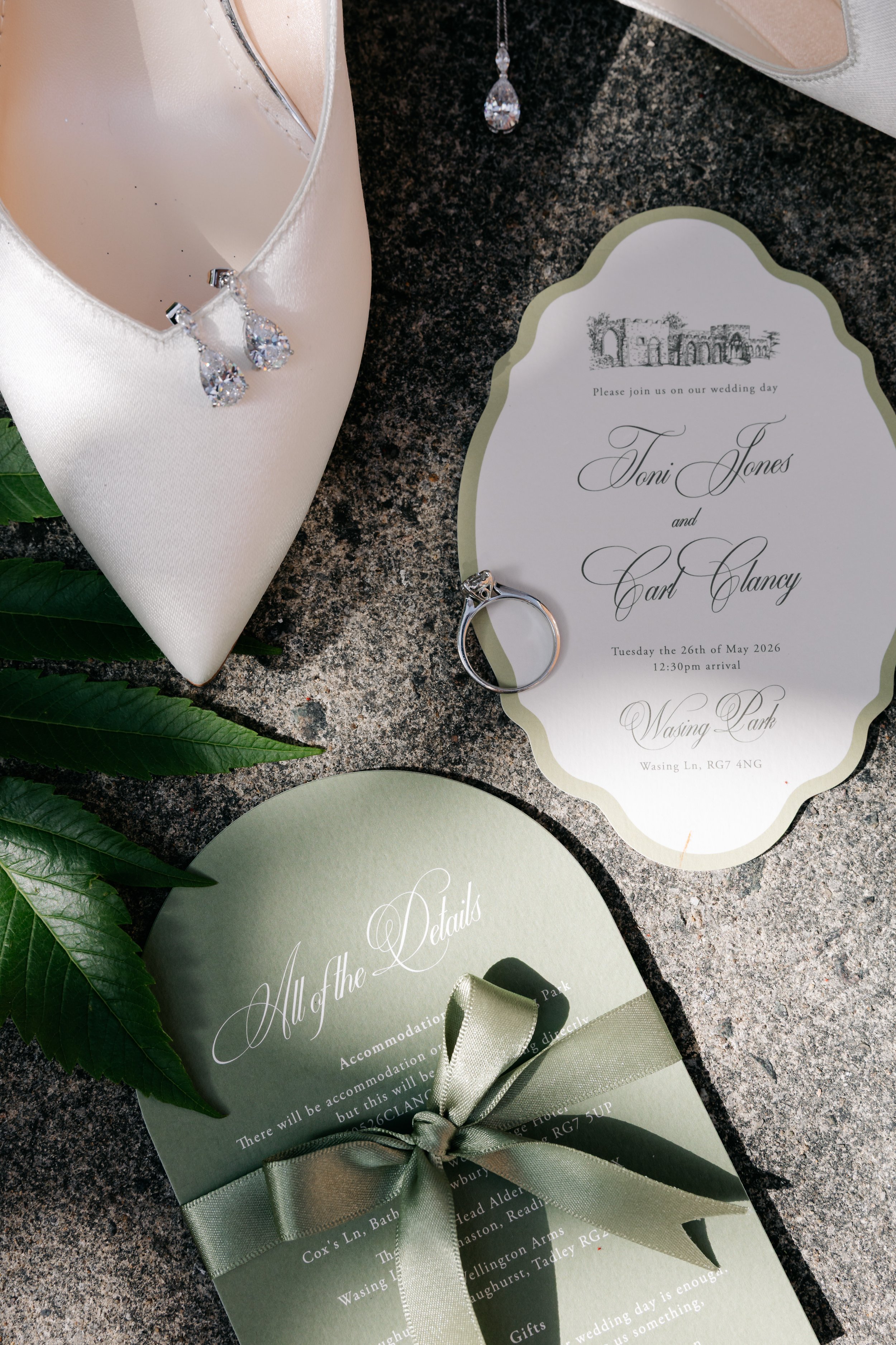 Wedding invitation with elegant text, a pair of diamond earrings on a satin cushion, and a wedding ring on a black granite surface.