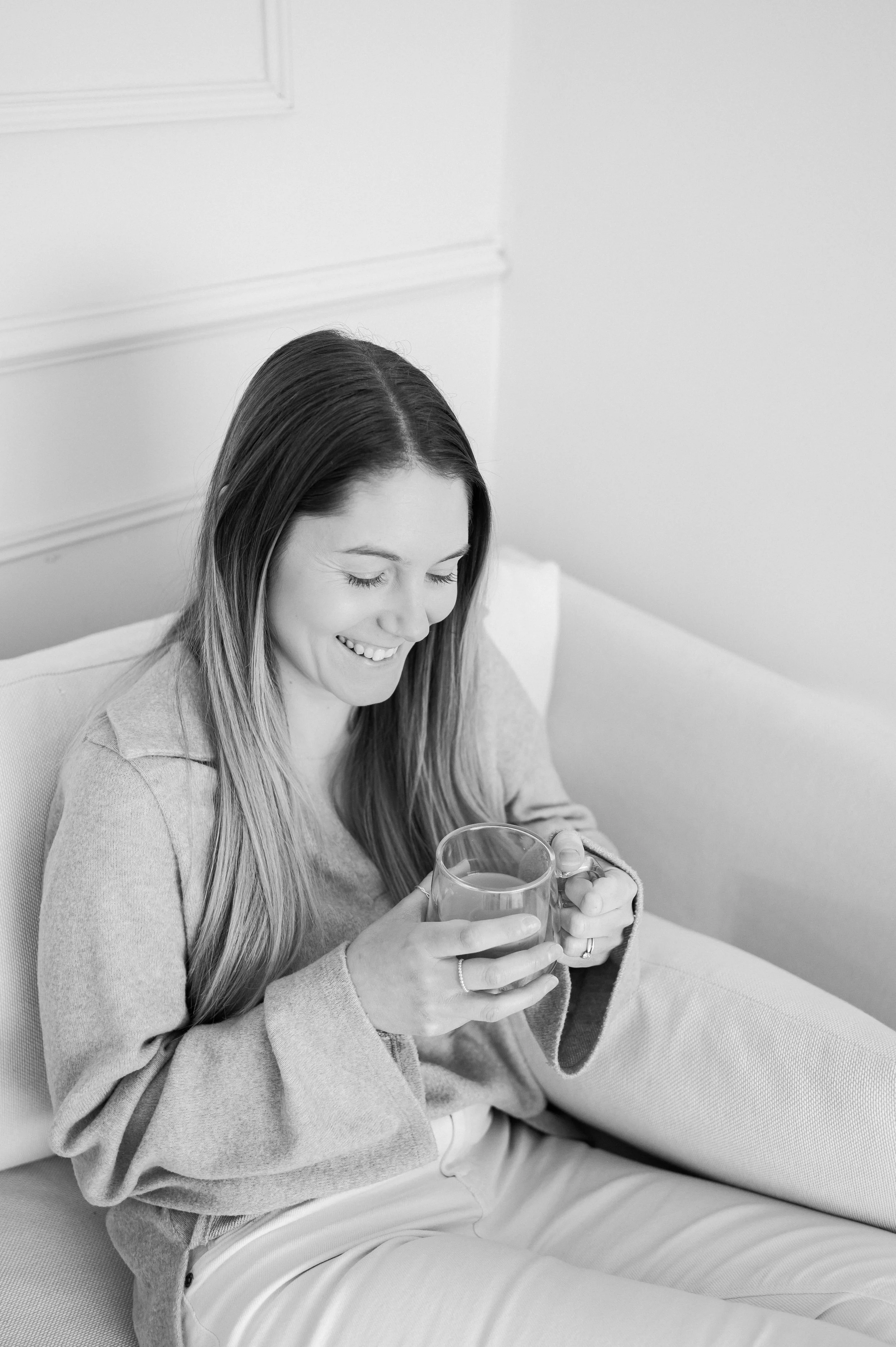 A woman with long hair sitting on a sofa, smiling, holding a glass mug, in a bright room.