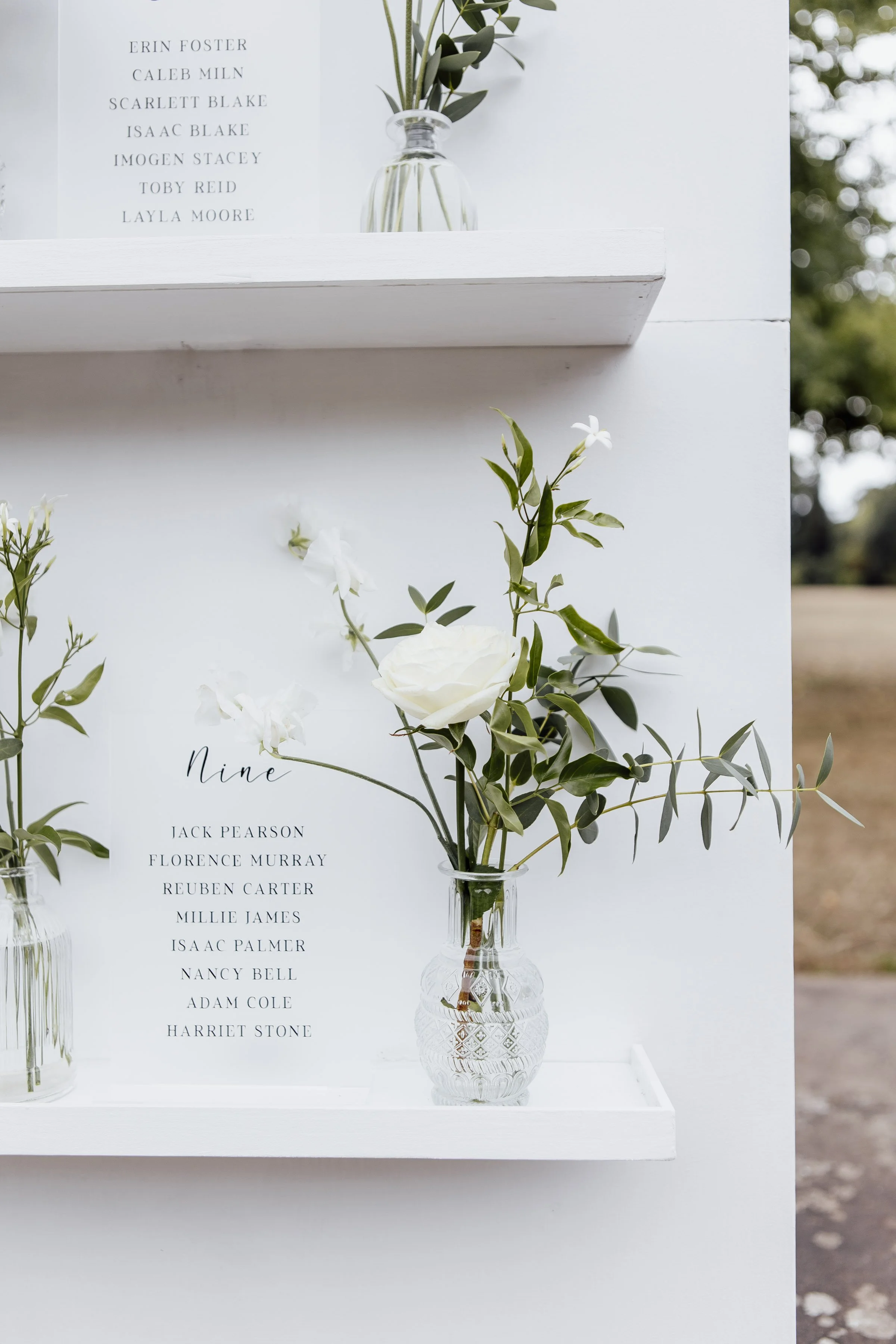 A white floral arrangement in a clear, patterned glass vase with green leaves and white flowers, placed on a white shelf. Behind it, a white sign with black text lists names under the heading 'Nine.' In the background, a white board displays more names in black text, and a glass vase with greenery sits atop the board. The scene appears to be part of an outdoor event decoration.
