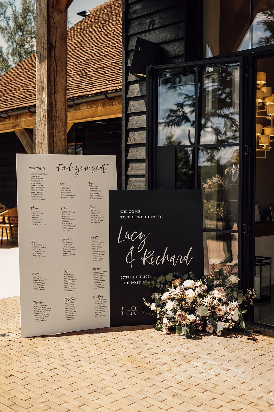 Wedding event sign with guest seating chart, wedding welcome sign for Lucy and Richard, date 27th July 2024, at The Post, decorated with a floral arrangement, outside of a rustic venue building with brick courtyard and black exterior walls.