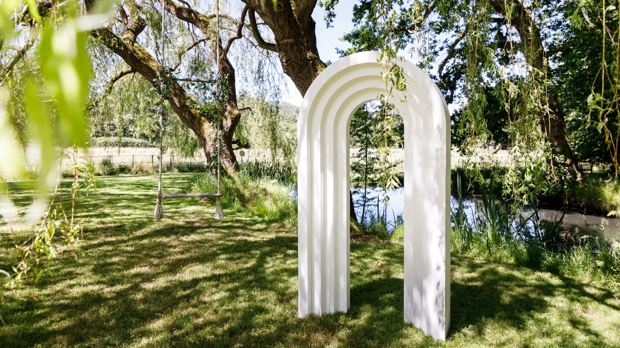 White decorative archway in a lush green park with trees, grass, and a pond in the background.