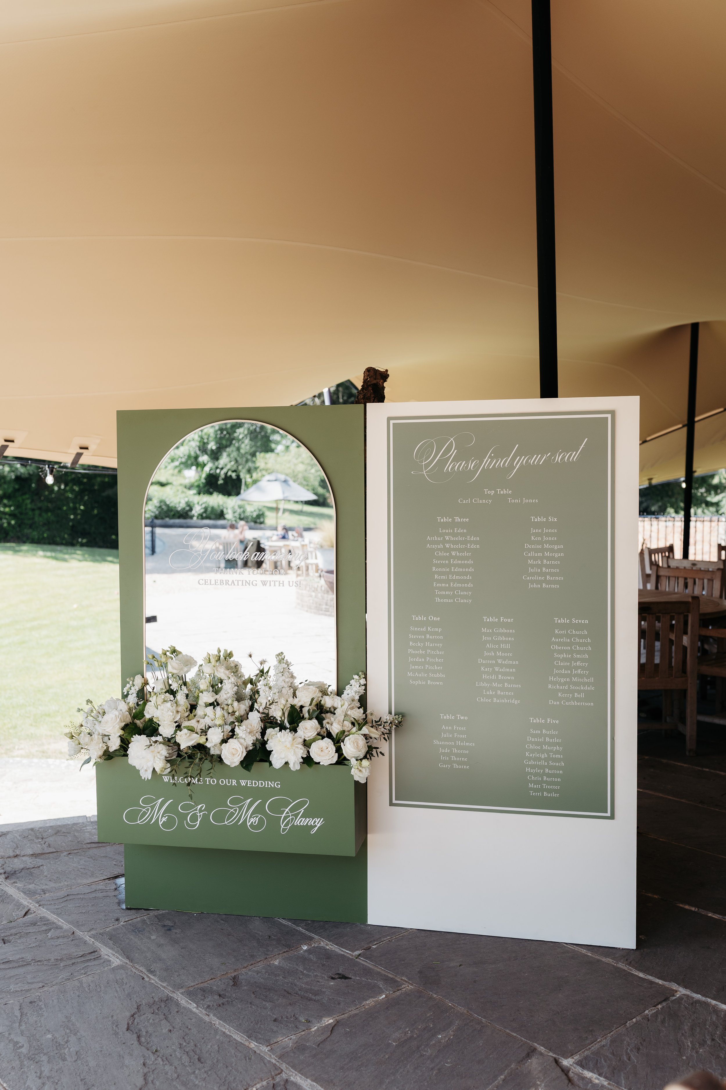 Wedding seating chart with a flower arrangement, set up outdoors under a canopy, with grass, trees, and an umbrella in the background.