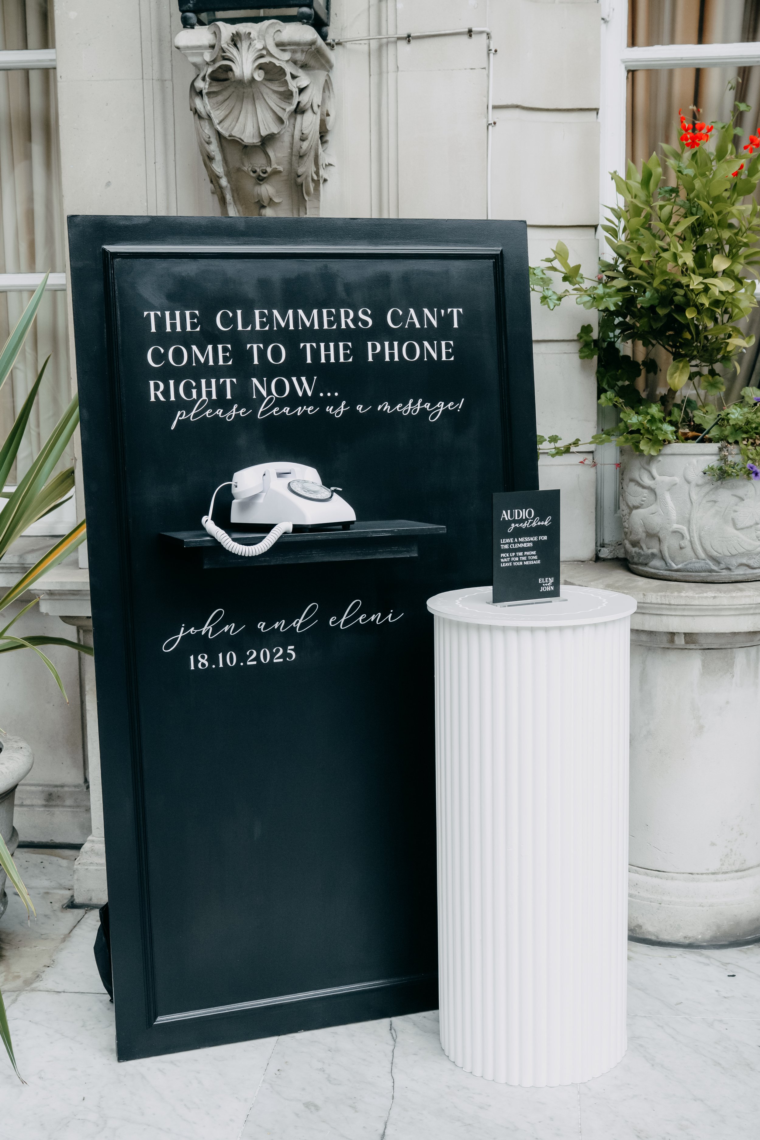 A black chalkboard with white text that reads, 'The Clemmers can't come to the phone right now... please leave us a message!' with a white telephone illustration and the names 'john and eleni' along with the date '18.10.2025'. A small black sign on a white pedestal provides instructions to leave a message for the couple.