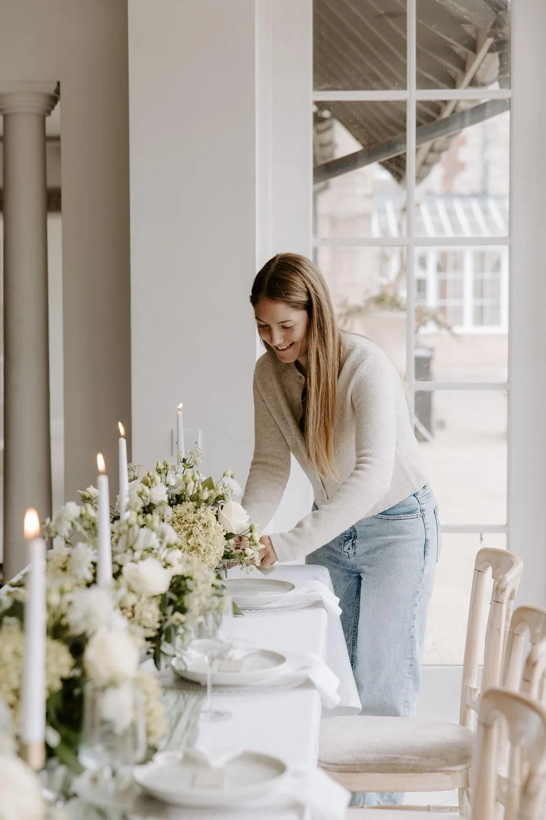 A young woman decorating a dining table with white flowers for an event, smiling as she arranges the table.