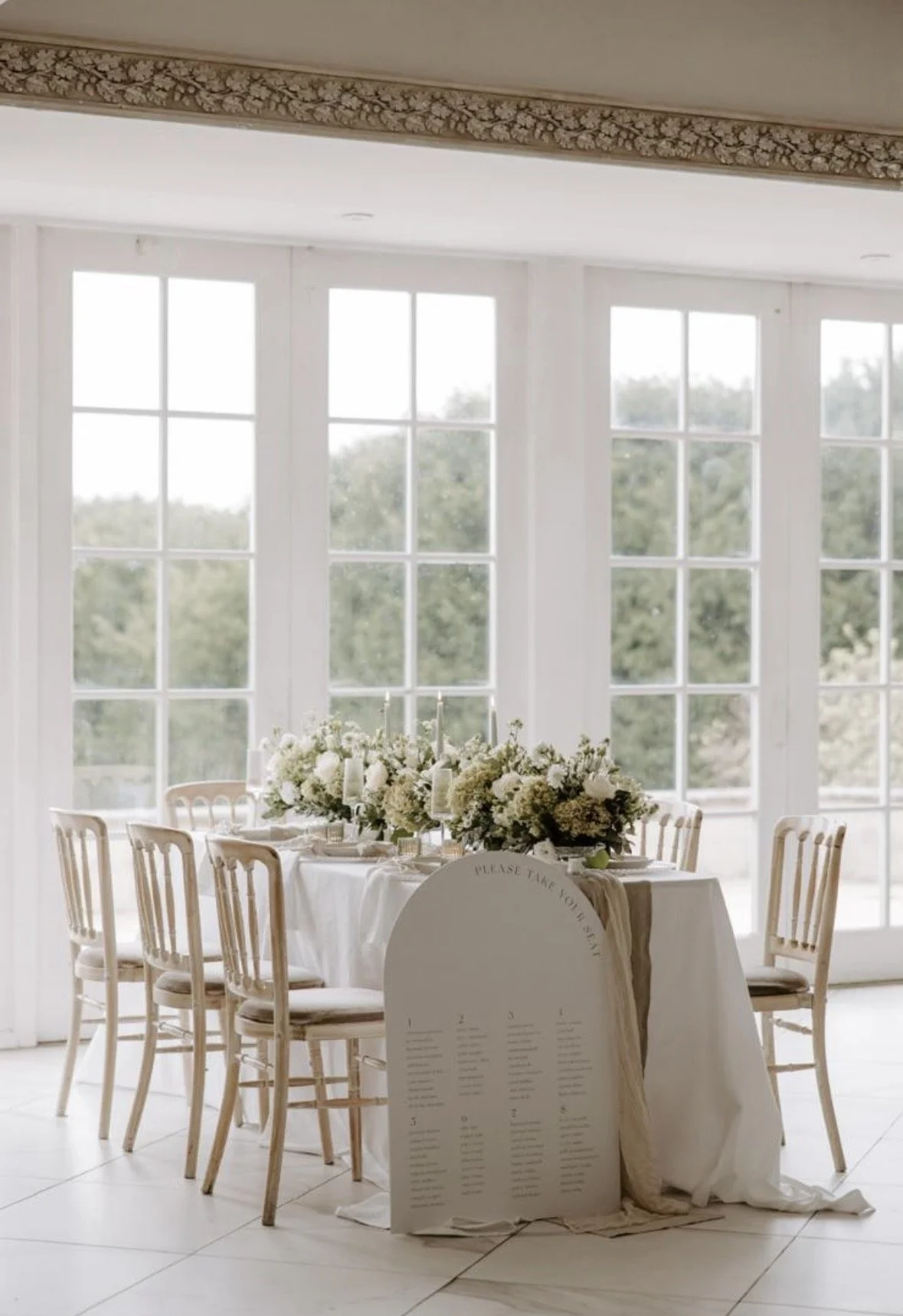 A round table decorated with a floral arrangement, candles, and tableware, situated near large windows with a trees view in the background. A seating chart is placed in front of the table.
