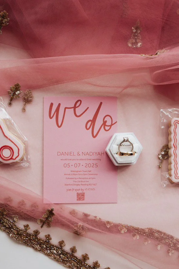 Pink wedding invitation card with gold text that says 'we do', accompanied by wedding rings in a white box, decorated with pink tulle, gold jewelry, cookies, and other wedding decorations.