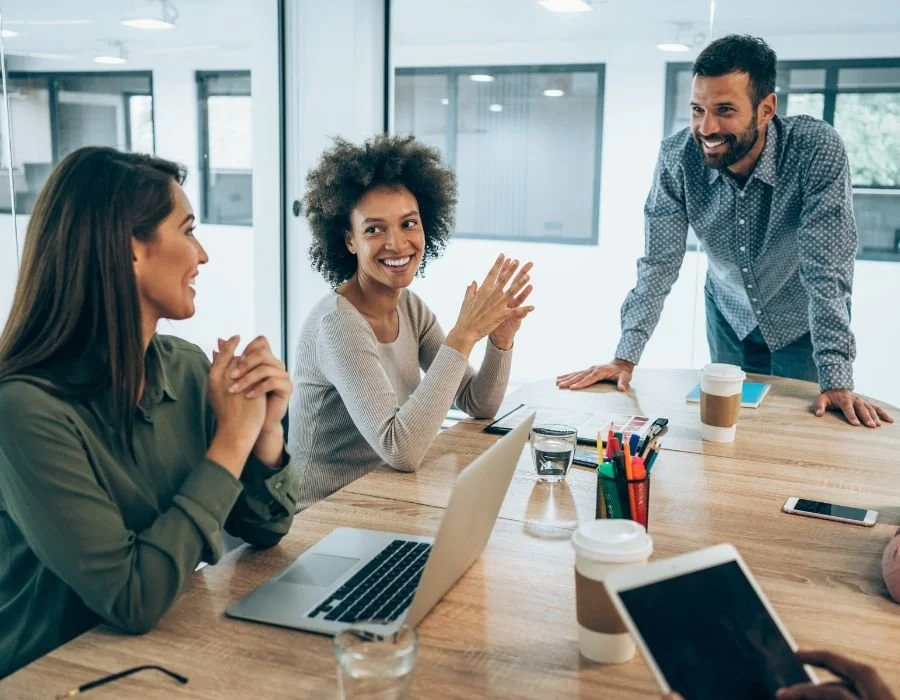 Seren Delta DIgital Transformation Services - Solution Discovery. Three people in a meeting room smiling and talking, with laptops, smartphones, coffee cups, and office supplies on the table.