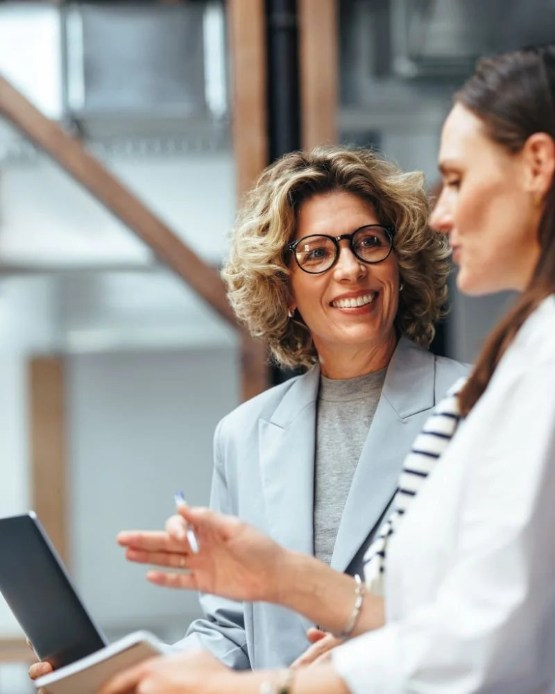 Seren Delta DIgital Transformation Services - Intelligent Architecture. Two women in a professional setting engaged in conversation, one holding a tablet and the other with a notepad, smiling and discussing.