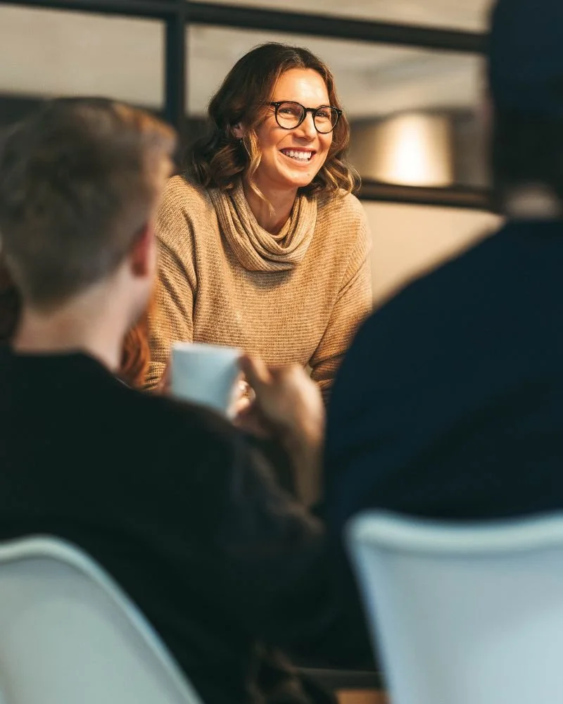 Seren Delta DIgital Transformation Services - A woman with glasses and wavy hair smiling during a conversation with two people in a casual setting, possibly a café or office.