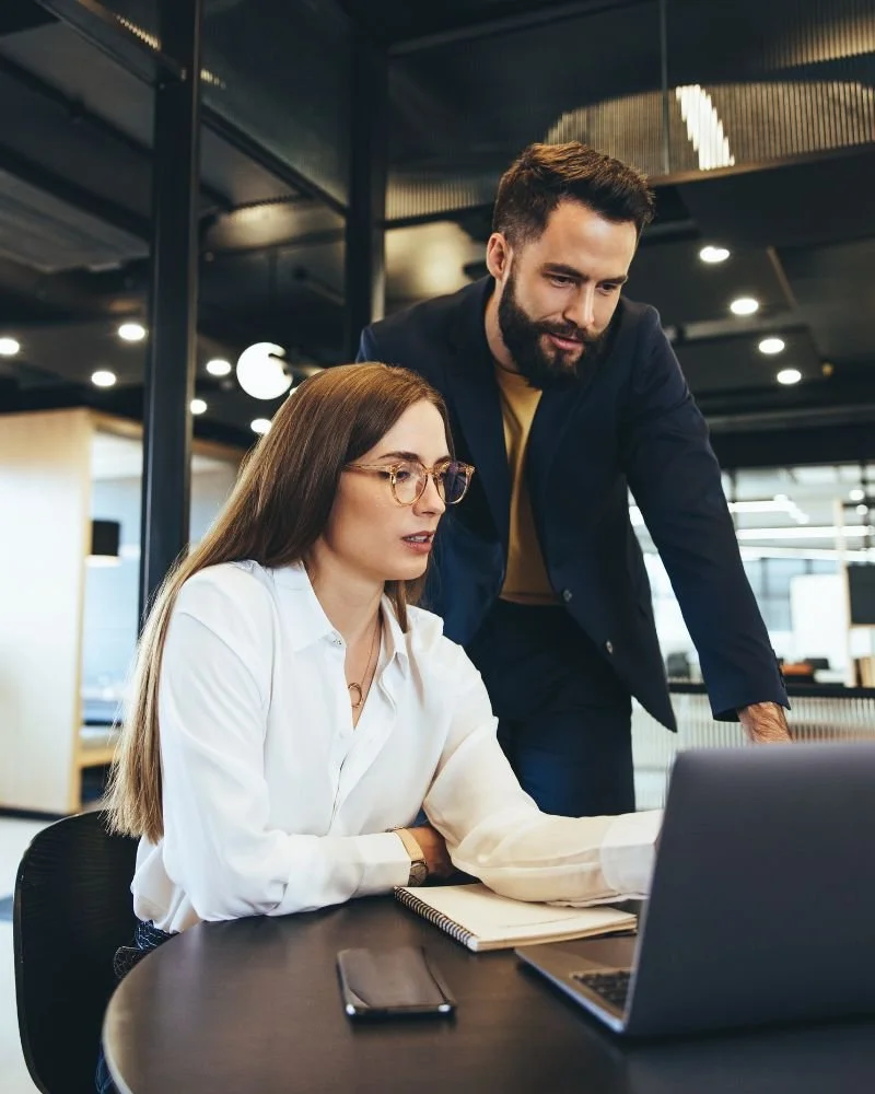 Seren Delta DIgital Transformation Services - Contact Us. A woman with glasses working on a laptop with a man standing and leaning over to look at the screen, in a modern office setting.