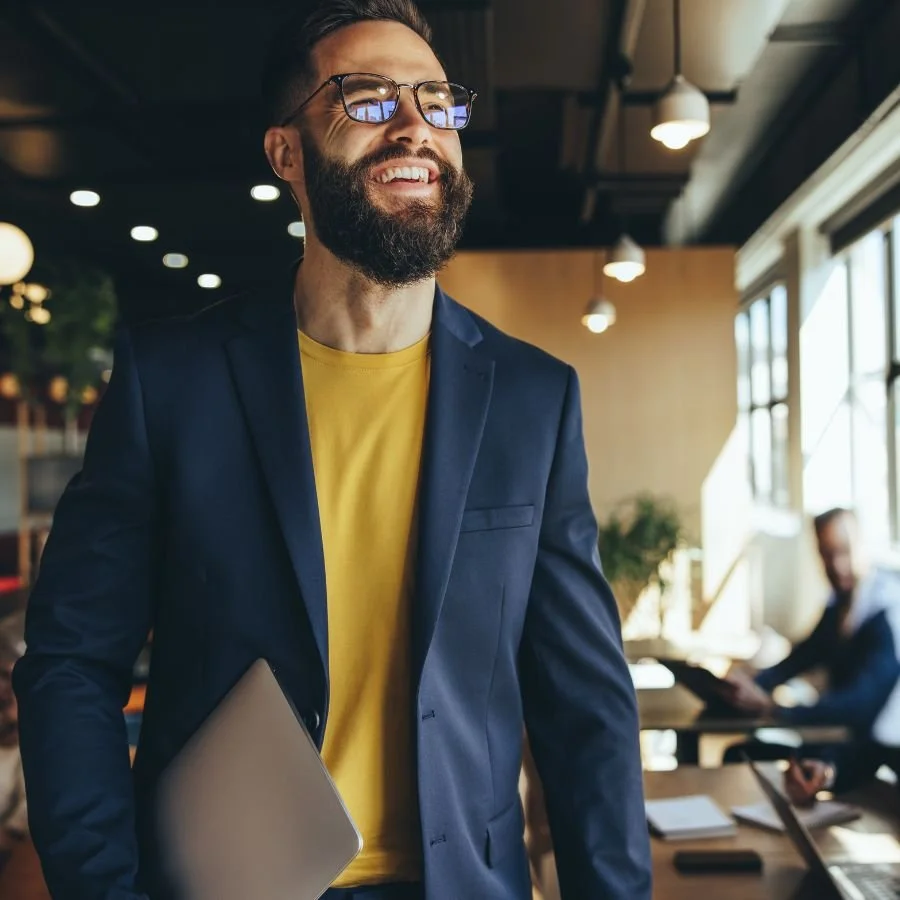 Seren Delta DIgital Transformation Services - About Us. A cheerful man with glasses and a beard holding a laptop in a modern office or coworking space.