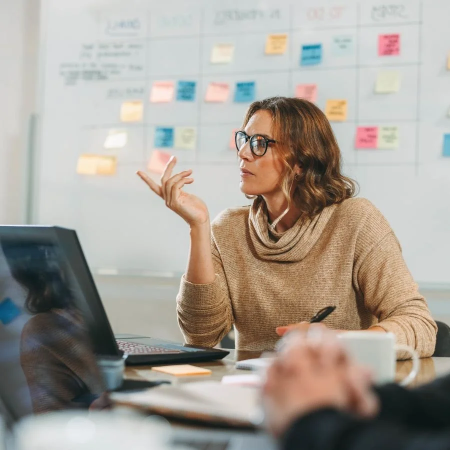Seren Delta DIgital Transformation Services - Solution Discovery. A woman wearing glasses and a beige sweater sitting at a desk, engaged in a discussion with a whiteboard covered in colorful sticky notes and handwritten notes in the background.