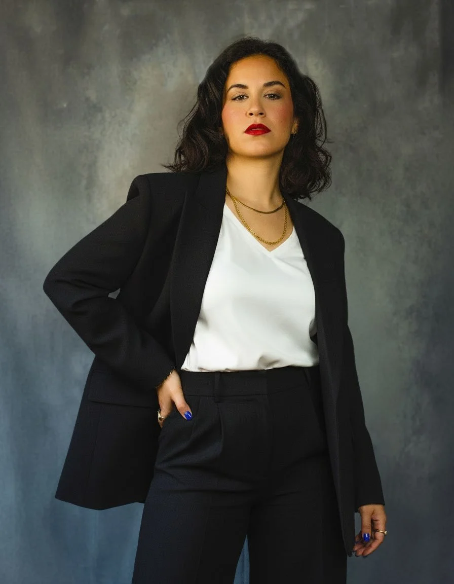 Businesswoman with dark wavy hair wearing a black blazer, white blouse, black pants, red lipstick, and gold jewelry, standing against a textured gray background.