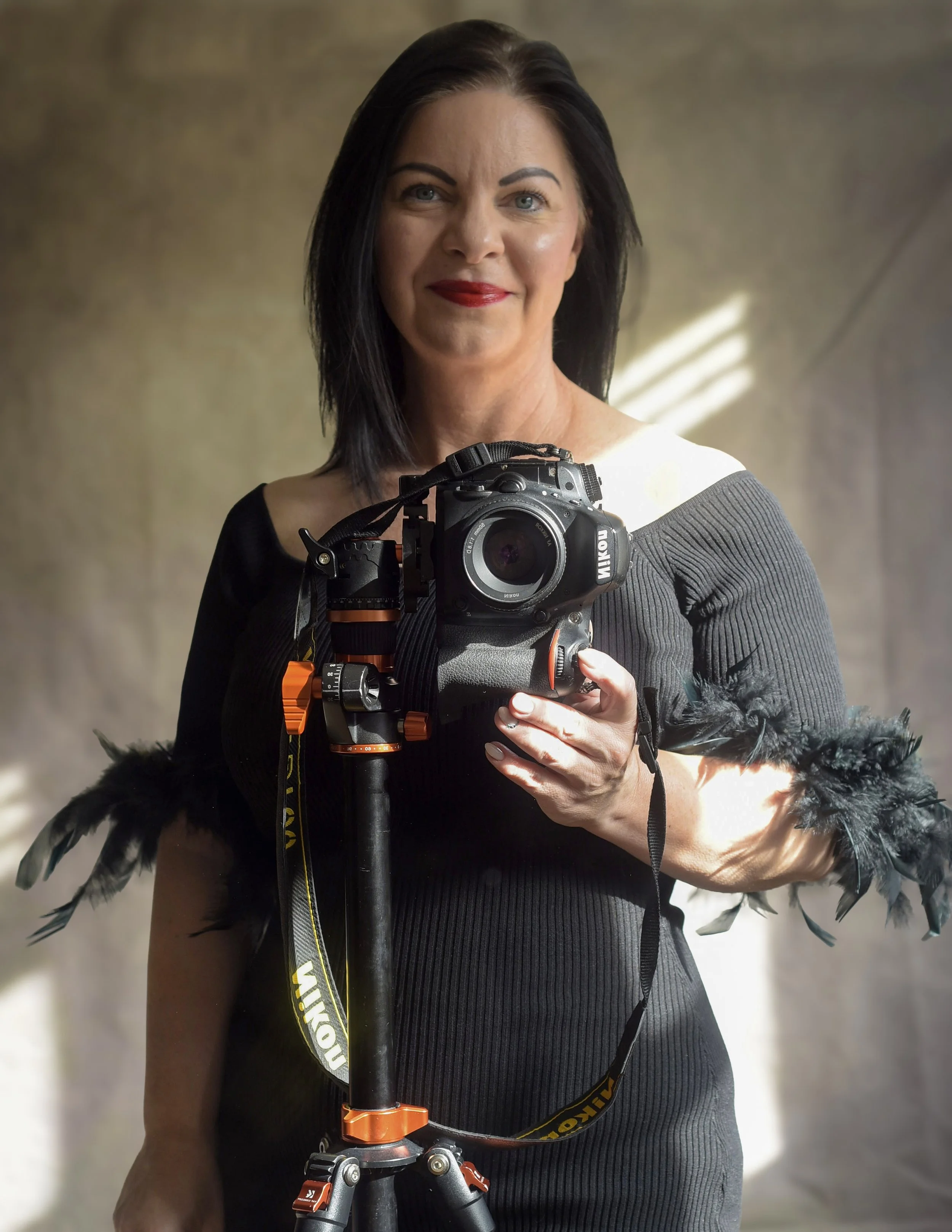 A woman with dark hair and red lipstick taking a self-portrait using a professional camera on a tripod.