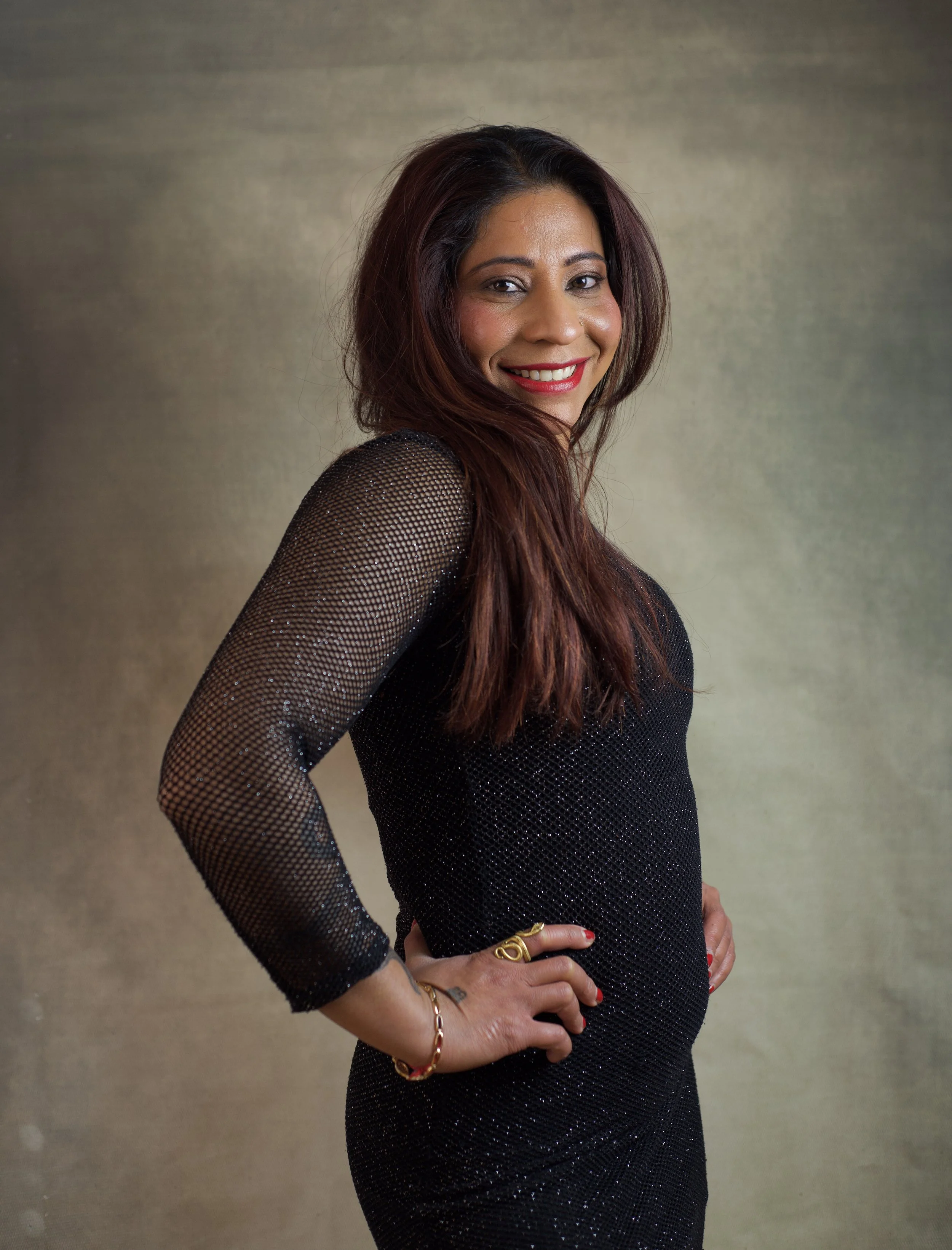 A woman with long dark hair smiling while standing sideways with hands on hips, wearing a black dress with sheer, sparkly sleeves against a neutral background.
