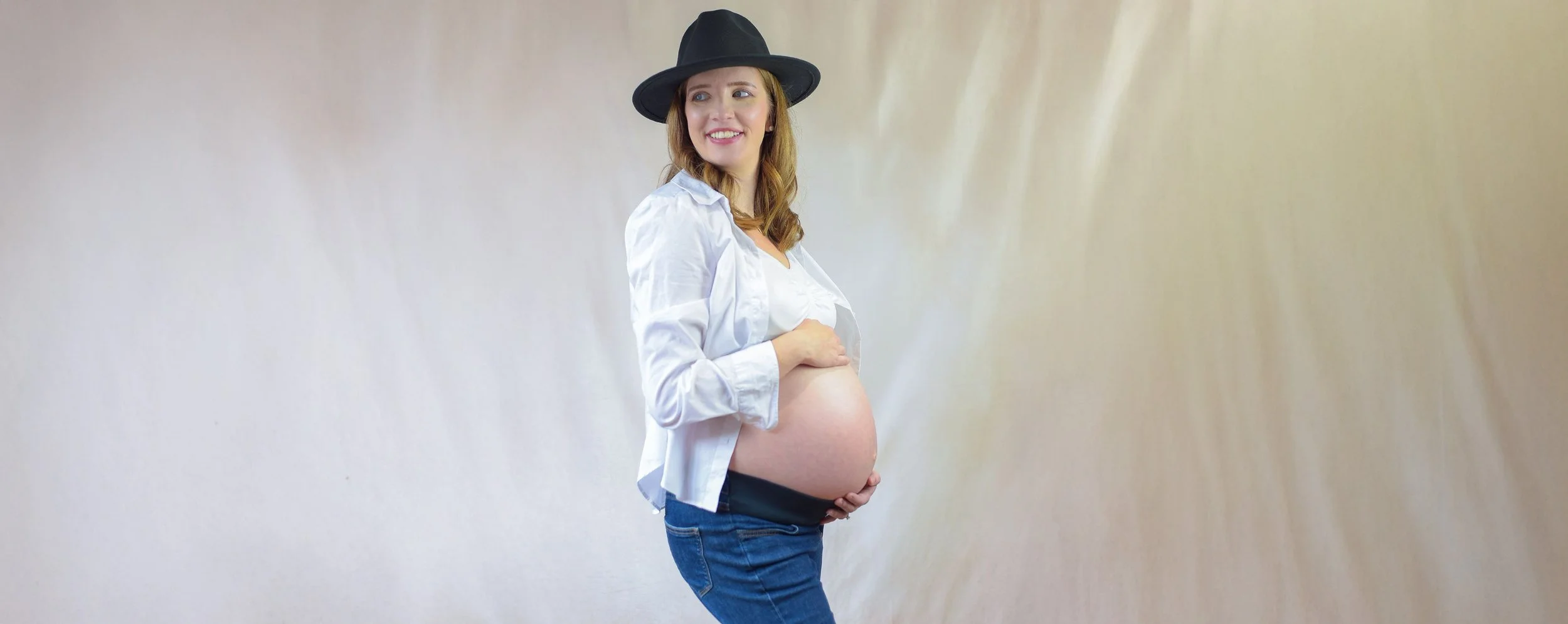 Pregnant woman wearing a black hat, white shirt, and jeans, holding her belly and smiling.