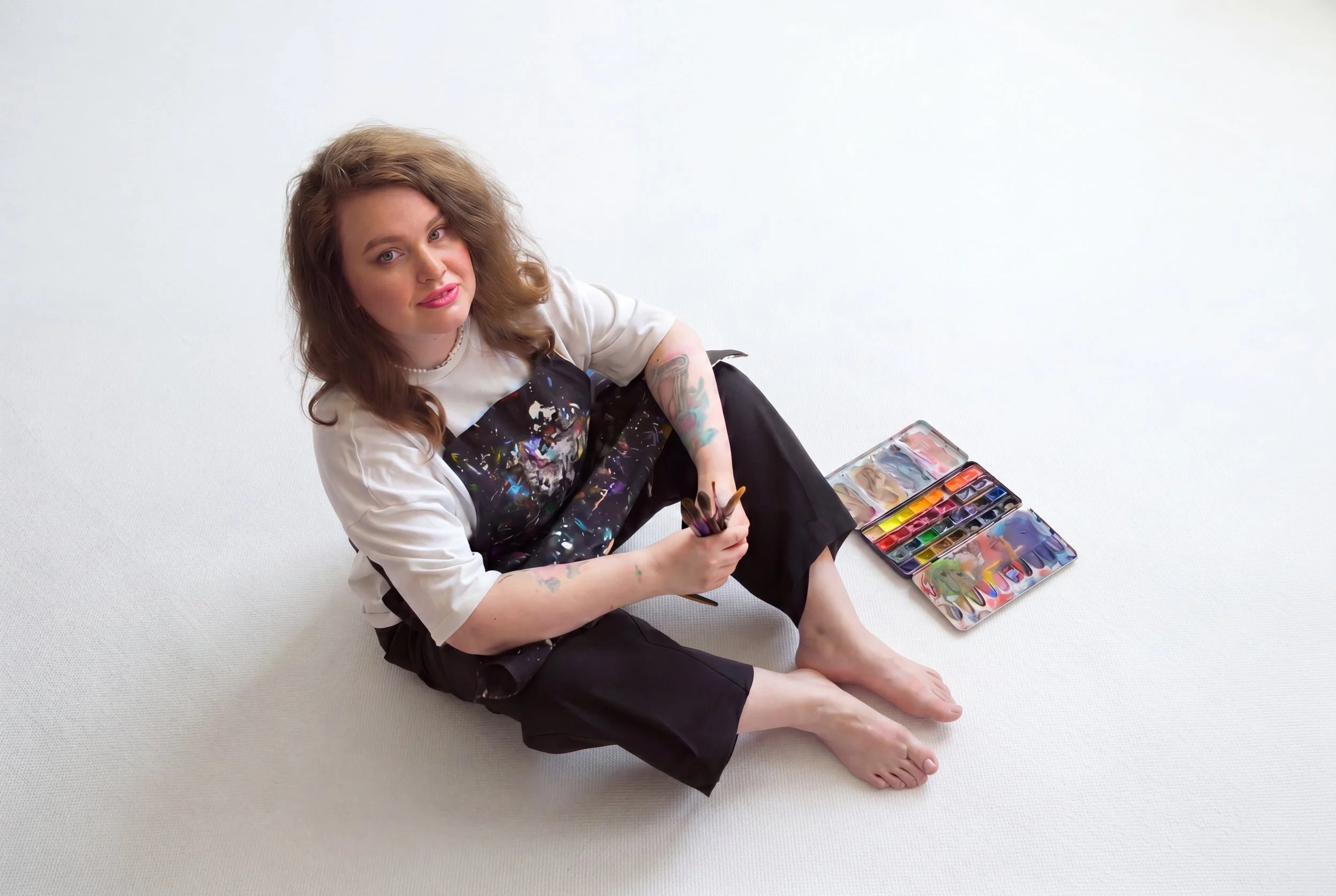 Young woman with tattoos sitting on the floor, holding a paintbrush, with colorful watercolor paints spread out beside her, against a plain white background.