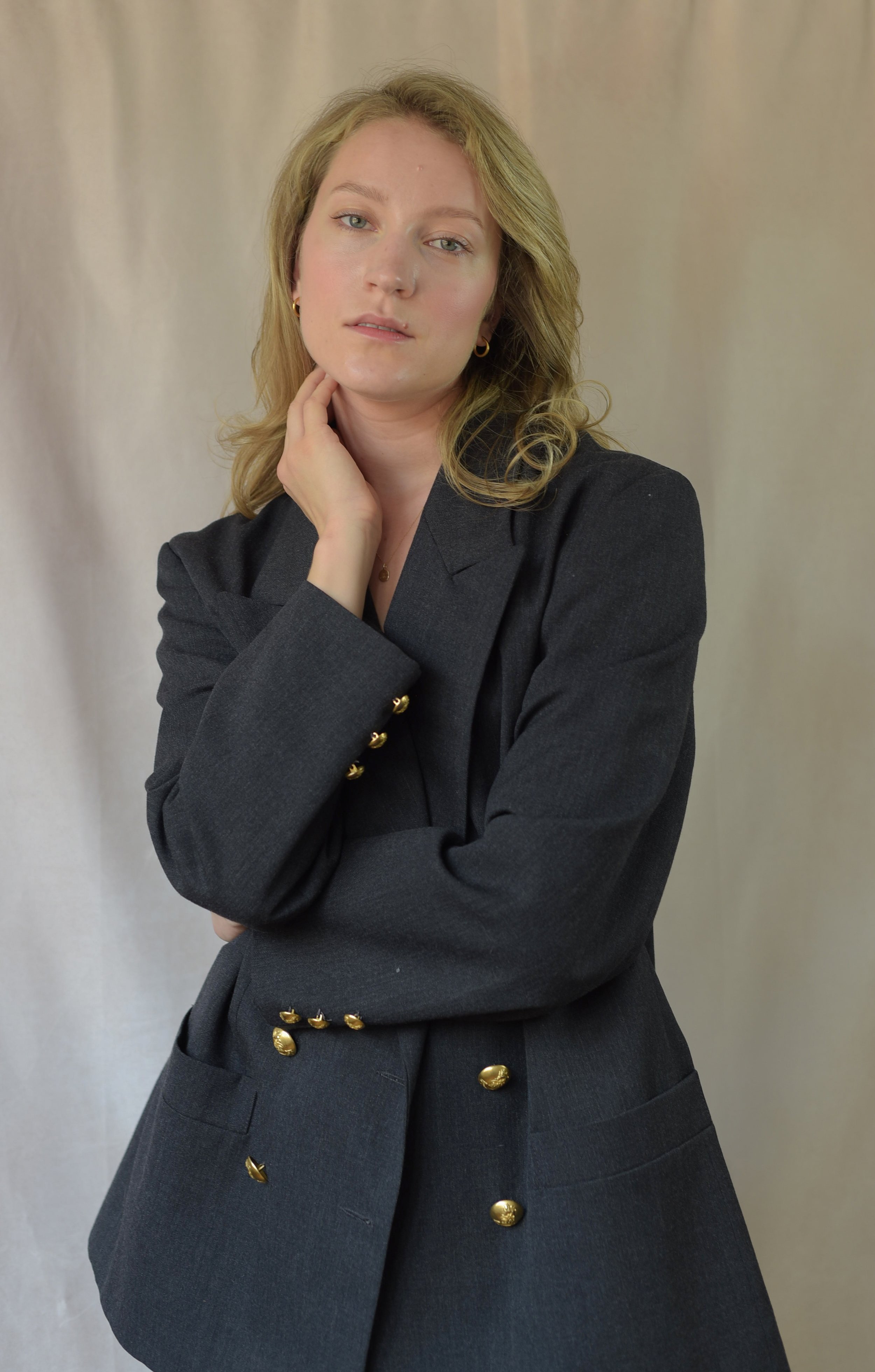 A woman with shoulder-length blonde hair in a dark blazer with gold buttons, posing with her hand near her neck against a plain backdrop.