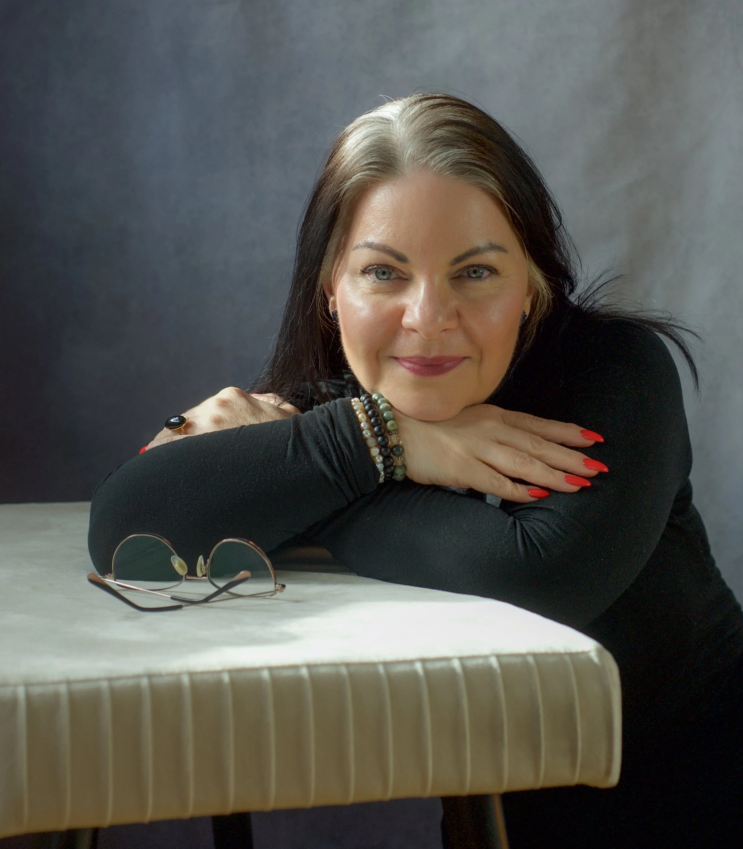A woman with gray and black hair, wearing multiple bracelets and a ring, resting her head on her crossed arms, smiling, with a pair of sunglasses placed on a beige textured surface in the foreground.