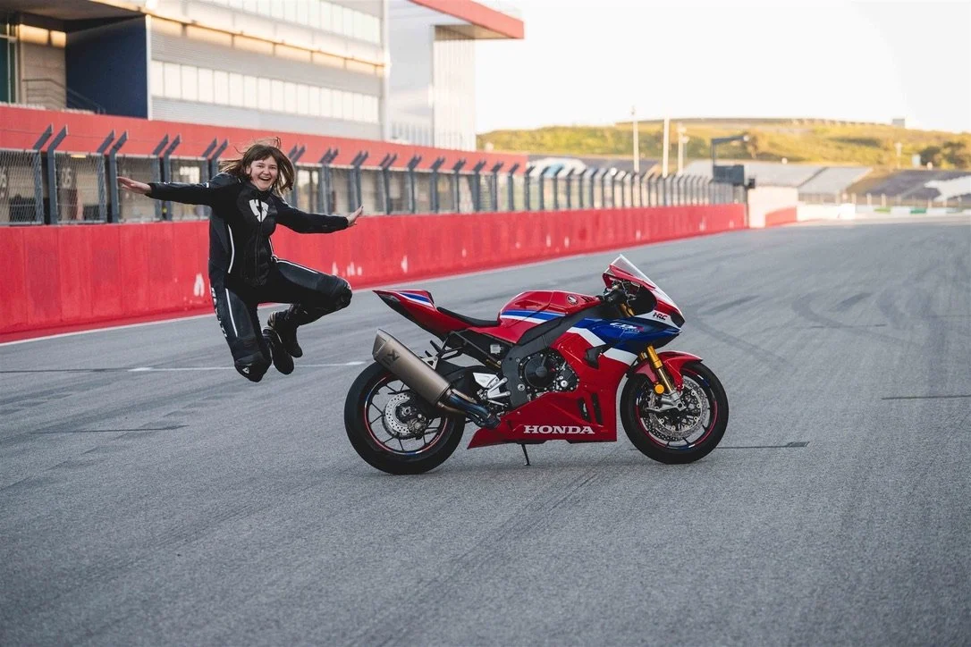 A person jumping in the air next to a red, white, and blue Honda sport motorcycle on a racetrack.