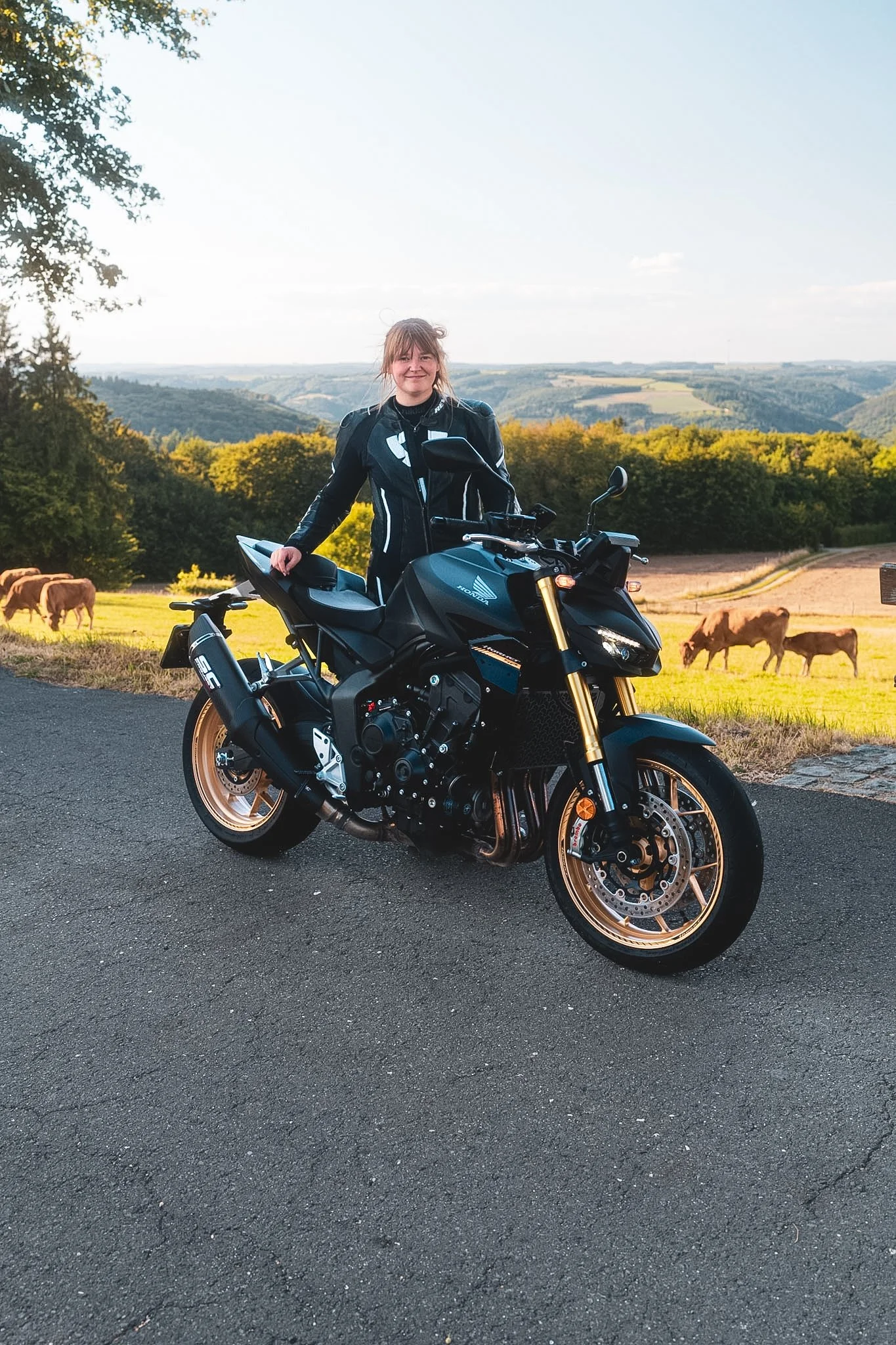 A woman in motorcycle riding gear standing next to a black motorcycle, Honda CB1000-SP on a rural road with a backdrop of green hills and grazing cows.