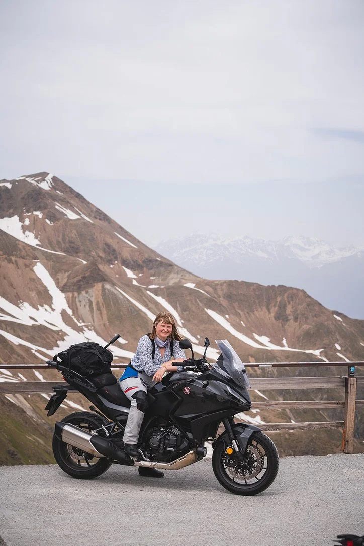 A woman sitting on a black motorcycle on a mountain road, with snow-capped mountain peaks in the background.