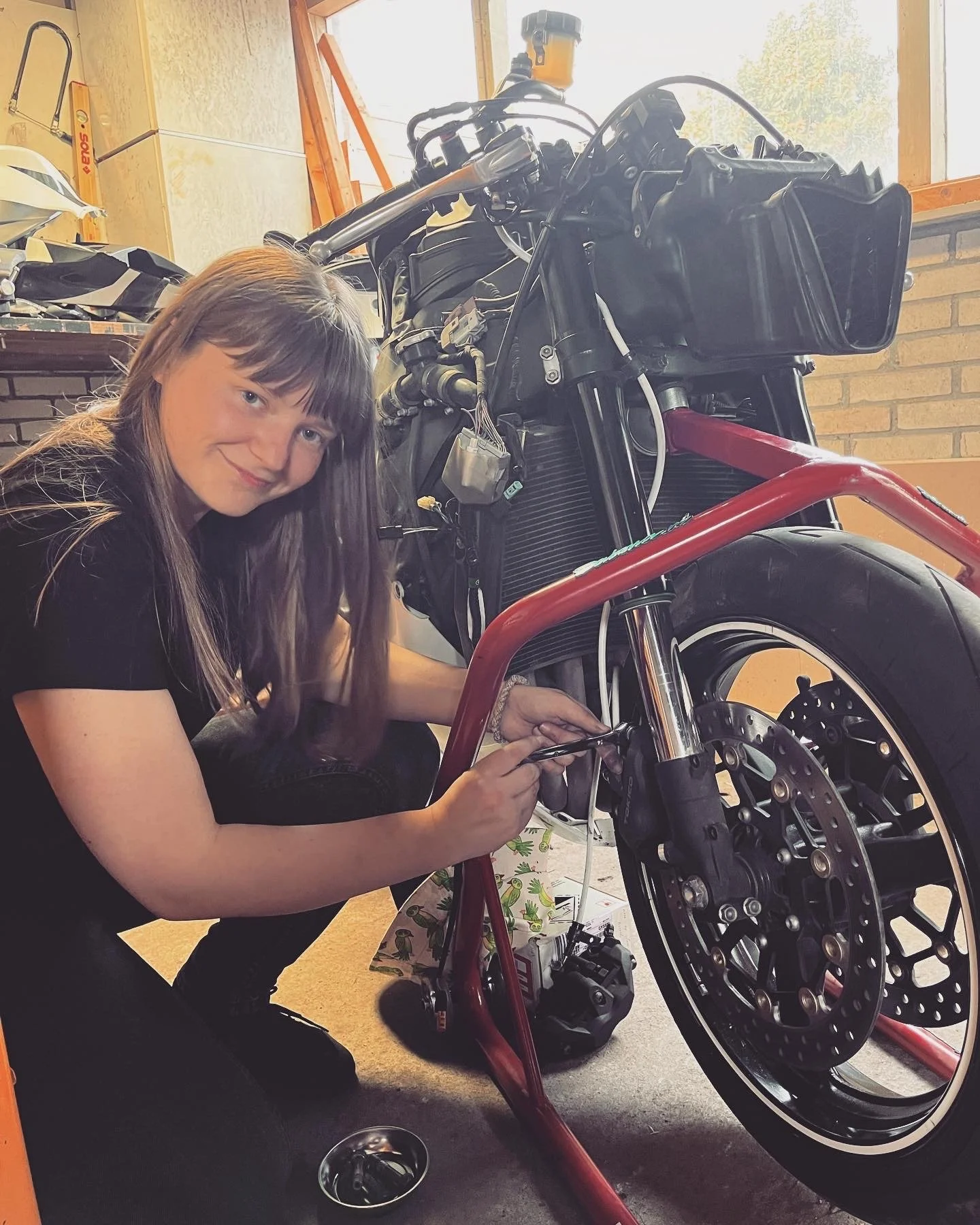 Meisje met lang haar heeft gereedschap vast en sleuteld aan een kale motor, ze lacht naar de camera