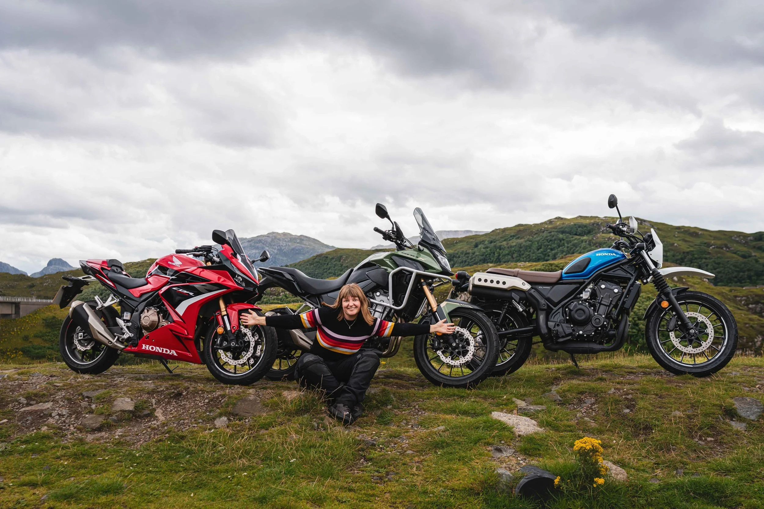 Girl sitting in front of 3 motorcycles, Honda 500CC engines, CBR500R, CB500X and CL500 in Scotland landscape