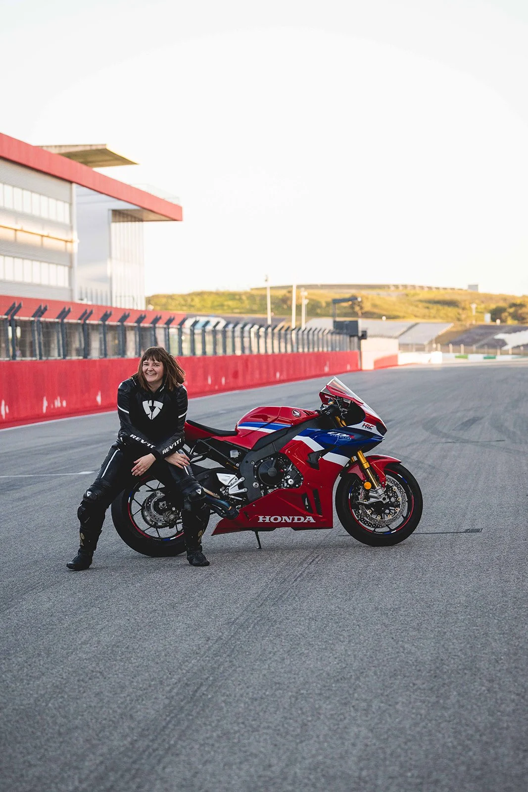 A woman in racing gear sitting on a red, white, and blue Honda motorcycle on a race track, smiling.