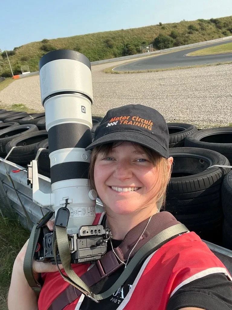 A young woman smiling at the camera while holding a large camera lens. She is outdoors at a racing circuit with stacked tires and a racetrack in the background. She is wearing a black cap with orange text that says 'Motor Circuit TRAINING' and a red racing vest.