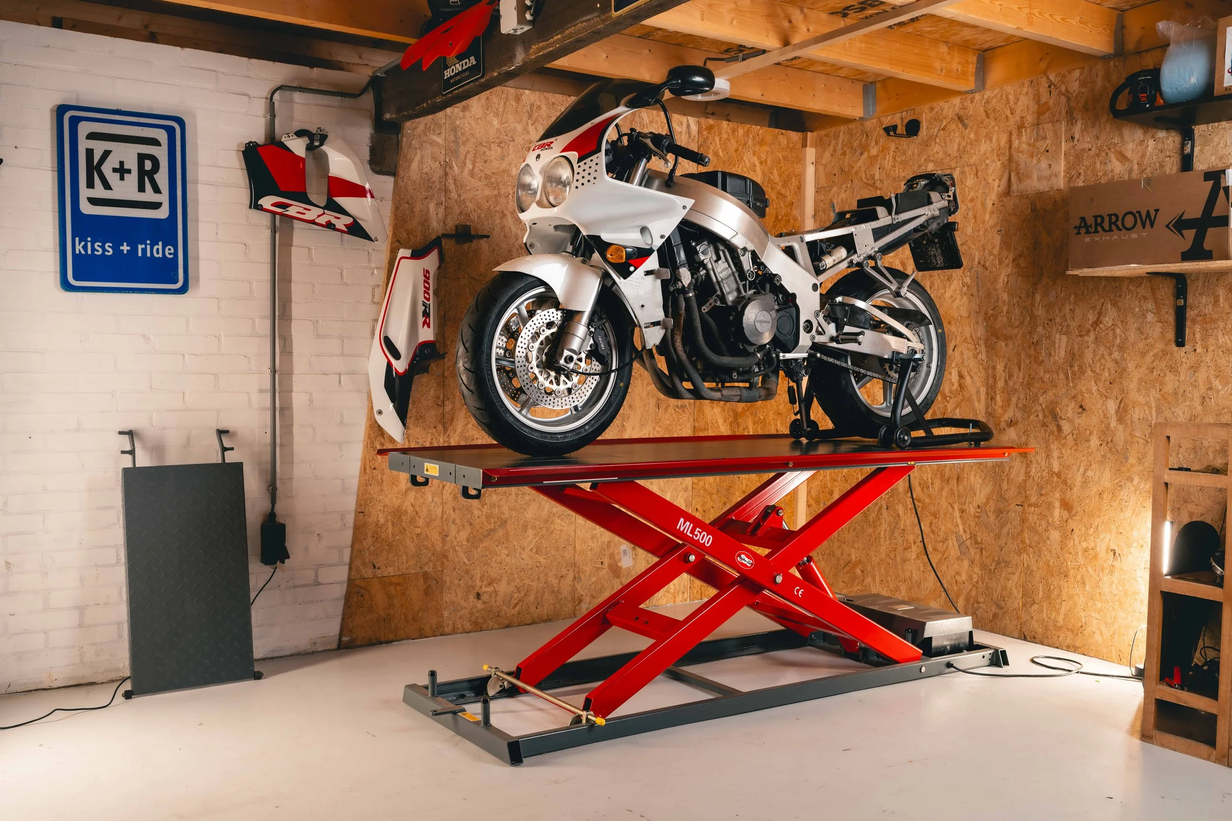 A motorcycle on a red repair lift inside a garage. The garage has a white brick wall with various motorcycle parts and signs, including a blue 'K+R kiss + ride' sign and Honda and Arrow Exhaust signs. The motorcycle is partially disassembled, with some parts removed and placed nearby.