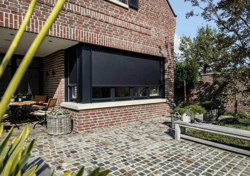 Ein Haus mit einer Terrasse, gepflastert mit Kieselsteinen, und einem Fenster mit geschlossenem schwarzen Sonnenschutz. Es gibt Gartenmöbel und Pflanzen im Garten.