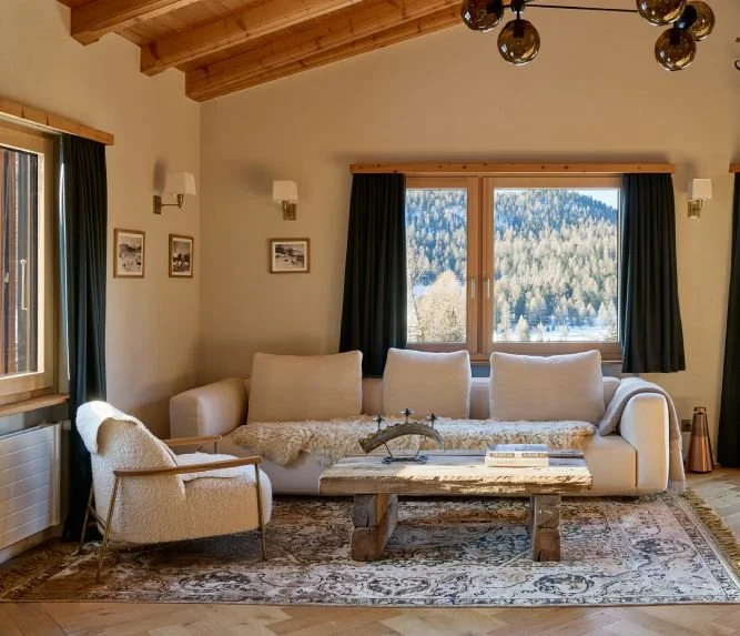 Ein gemütliches Wohnzimmer mit einem weißen Sofa und einem Armchair, einem rustikalen Holztisch, dunklen Vorhängen, einer Teppichläge und einem großen Holzfenstern mit Blick auf eine Alpenlandschaft.