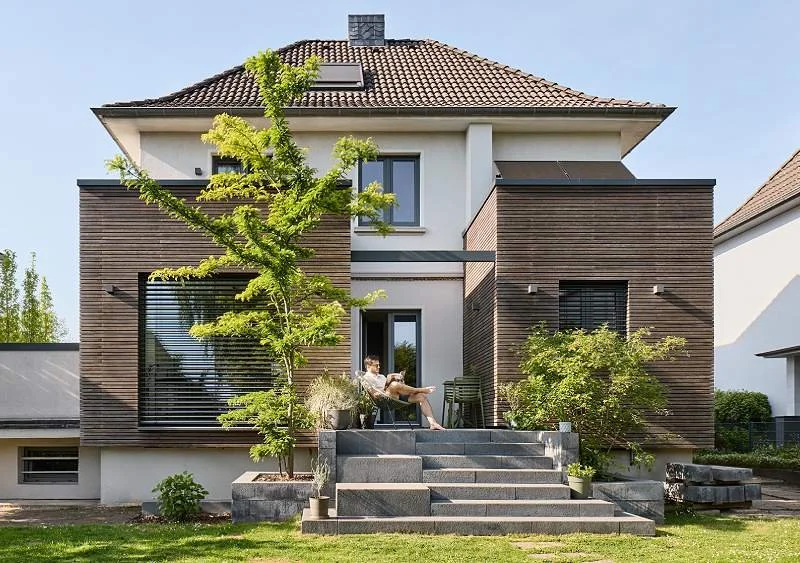 Modernes zweistöckiges Haus mit Holzverkleidung, große Fenster mit Raffstoren, Dach mit Dachfenster, vor dem Eingang sitzt eine Person auf den Stufen, umgeben von grünen Bäumen und Pflanzen.