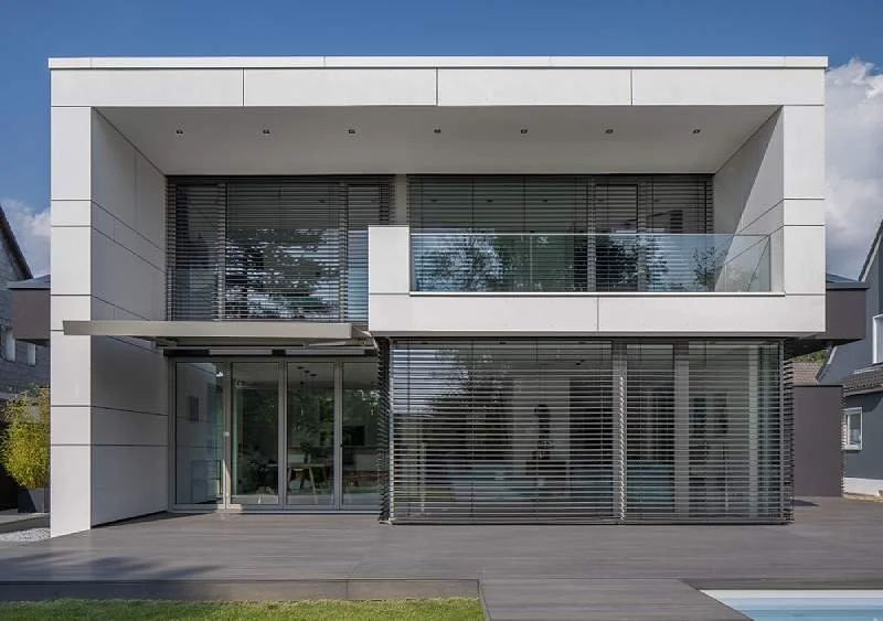 Modernes, zweistöckiges Haus mit großen Fenstern, Aluminium-Raffstoren, Glasbalustrade und einer Holzterrasse im Vordergrund.